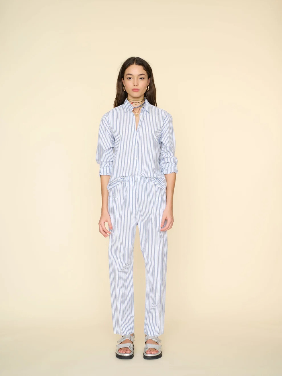 A woman in a striped jumpsuit and sandals stands against a pale yellow background.