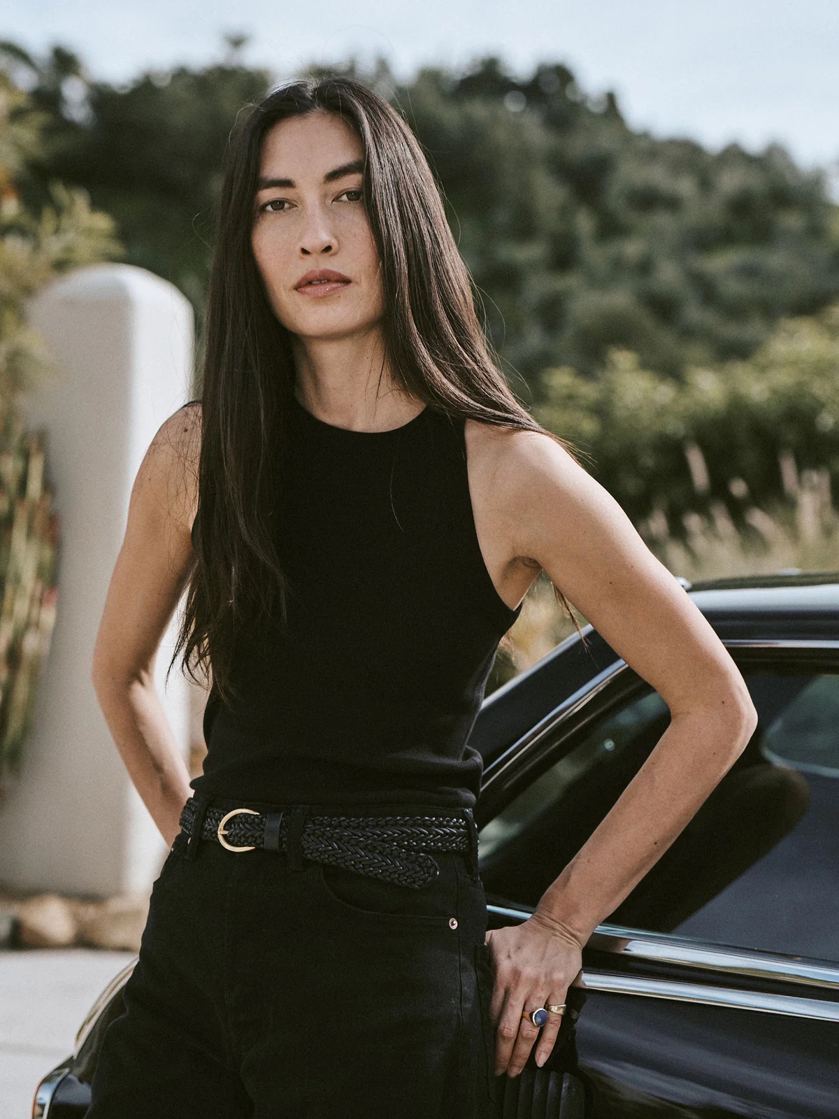 A woman in a black tank top and jeans stands confidently by her car, hands in pockets, against a natural backdrop.