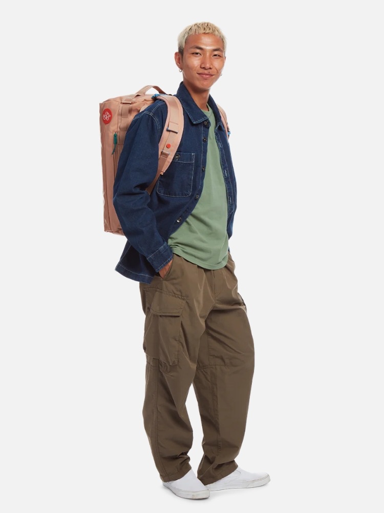 Young man with bleached hair, wearing a denim jacket, green shirt, and cargo pants, standing with a backpack, isolated on a white background.