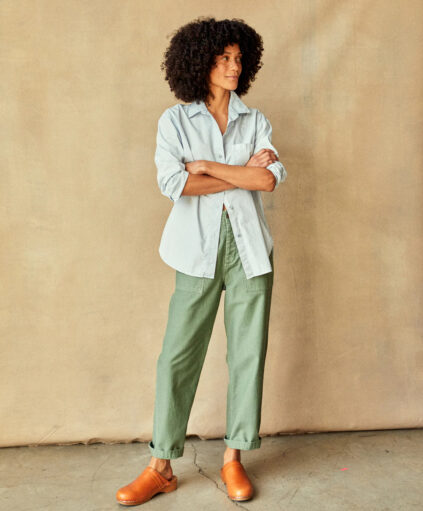 A person with curly hair standing against a beige background, wearing a casual blue shirt, green pants, and brown shoes, with arms crossed.