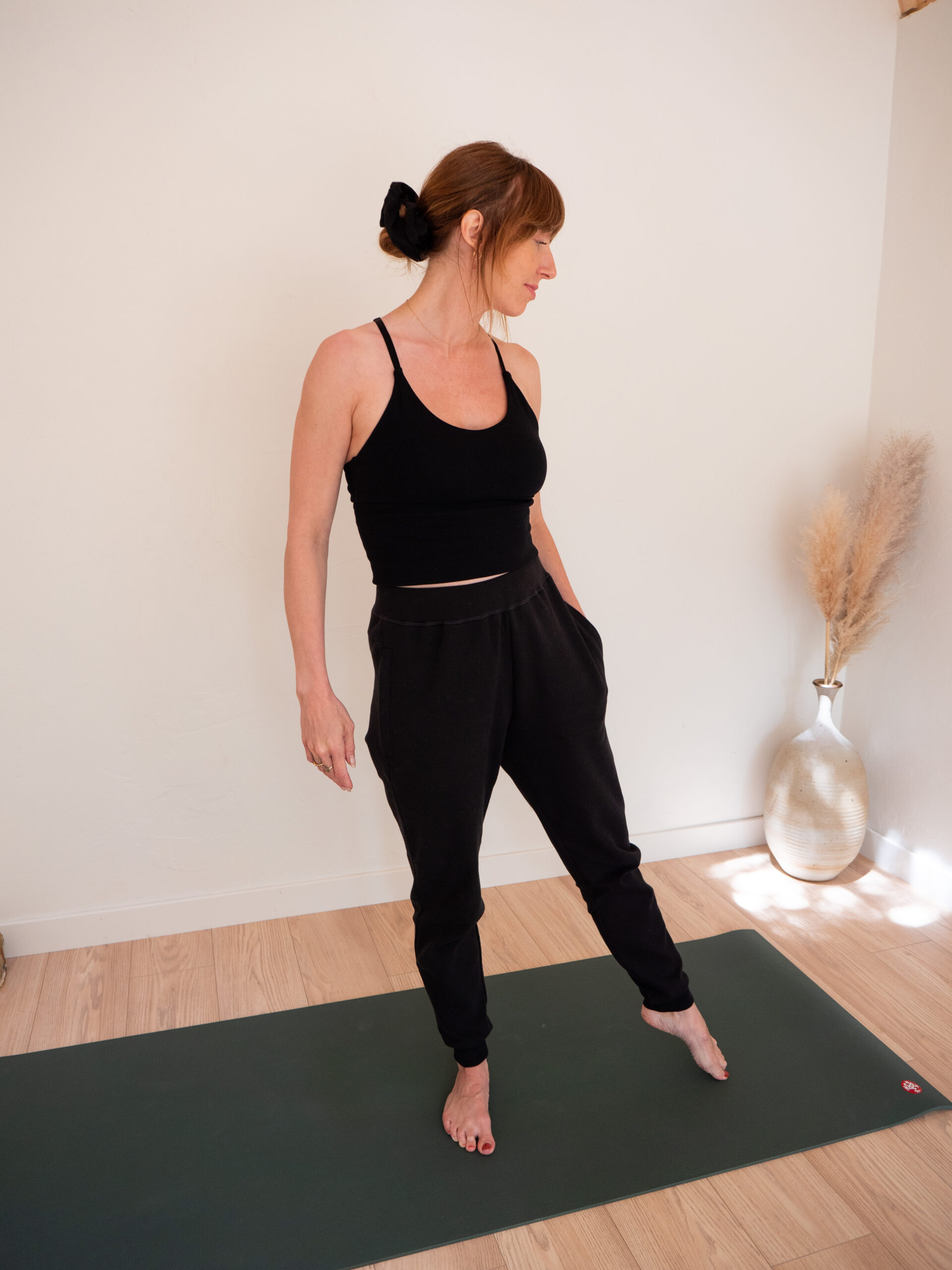 A person in a black tank top and black pants stands on a green yoga mat in a brightly lit room with a vase of dried plants in the background.