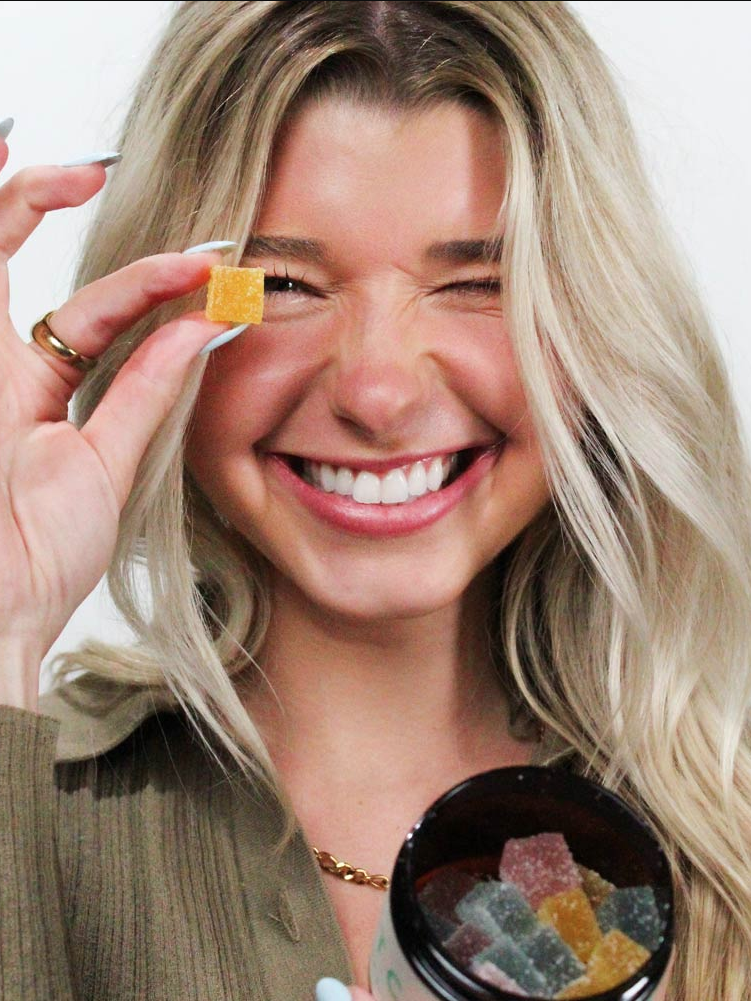 A woman with blond hair smiling widely, holding a gummy candy to her eye, and a jar of gummies in her other hand.