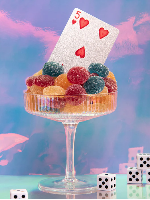 A glass filled with colorful gummy candies, topped with a glittery five of hearts card, surrounded by dice, against a pastel background.