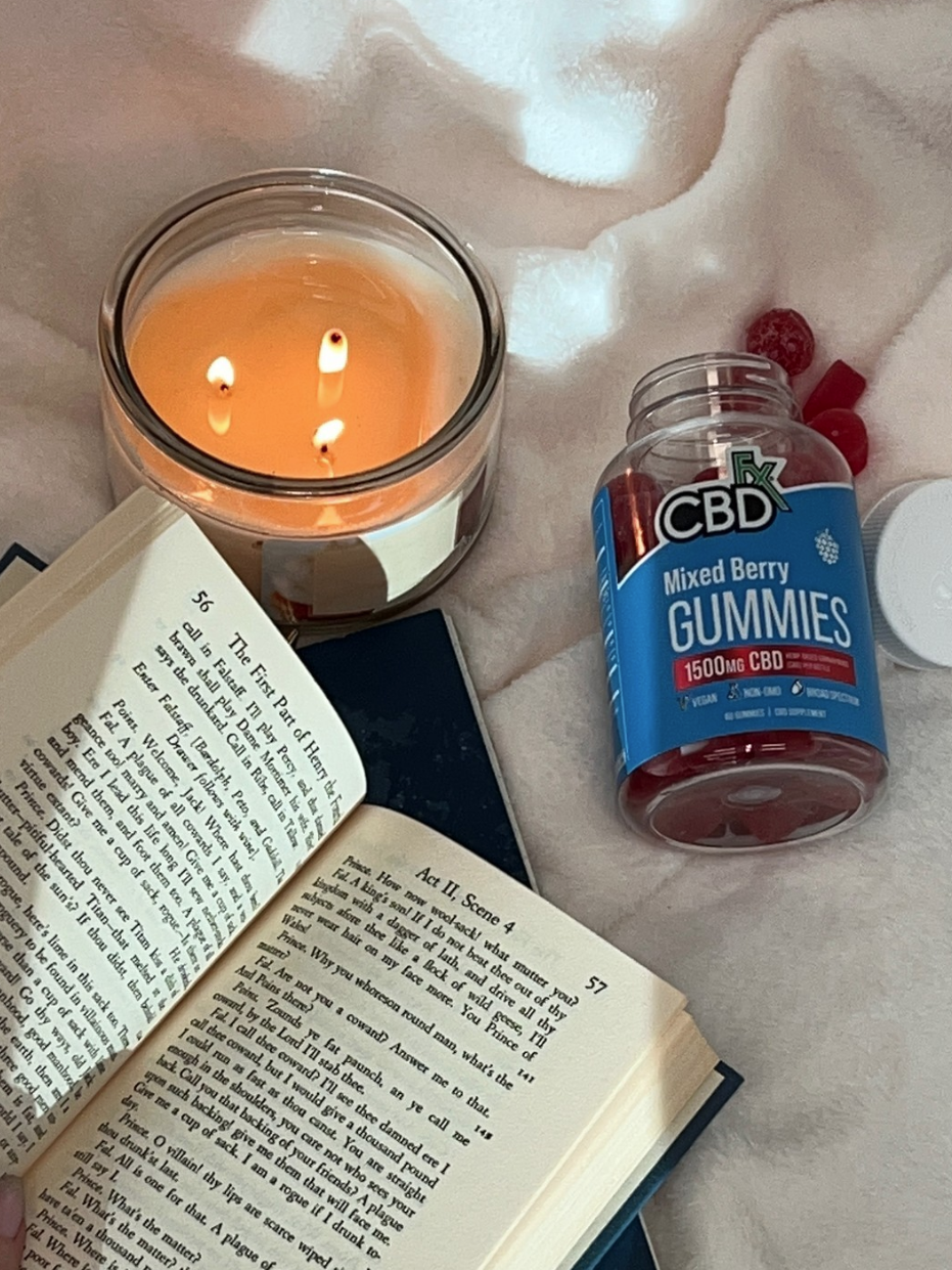 A person reading a book beside a lit candle and a bottle of cbd gummies on a soft, white blanket.