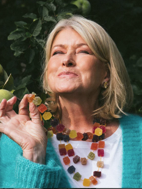A woman in a teal sweater smiles with closed eyes, holding colorful gummy candies in her hand, surrounded by green foliage.