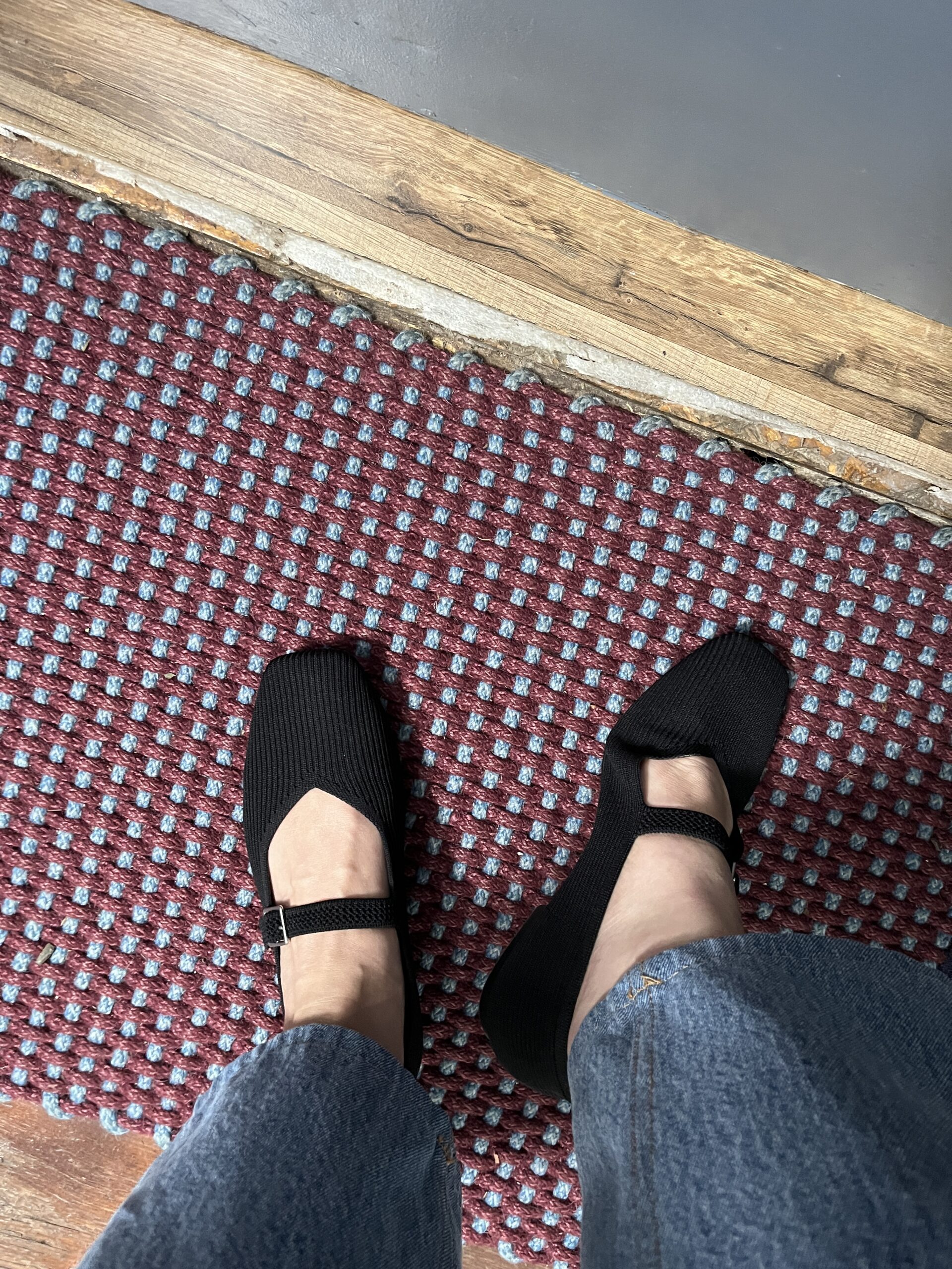 A person wearing black Mary Jane style shoes and blue jeans stands on a red and blue textured carpet near a wooden floor.