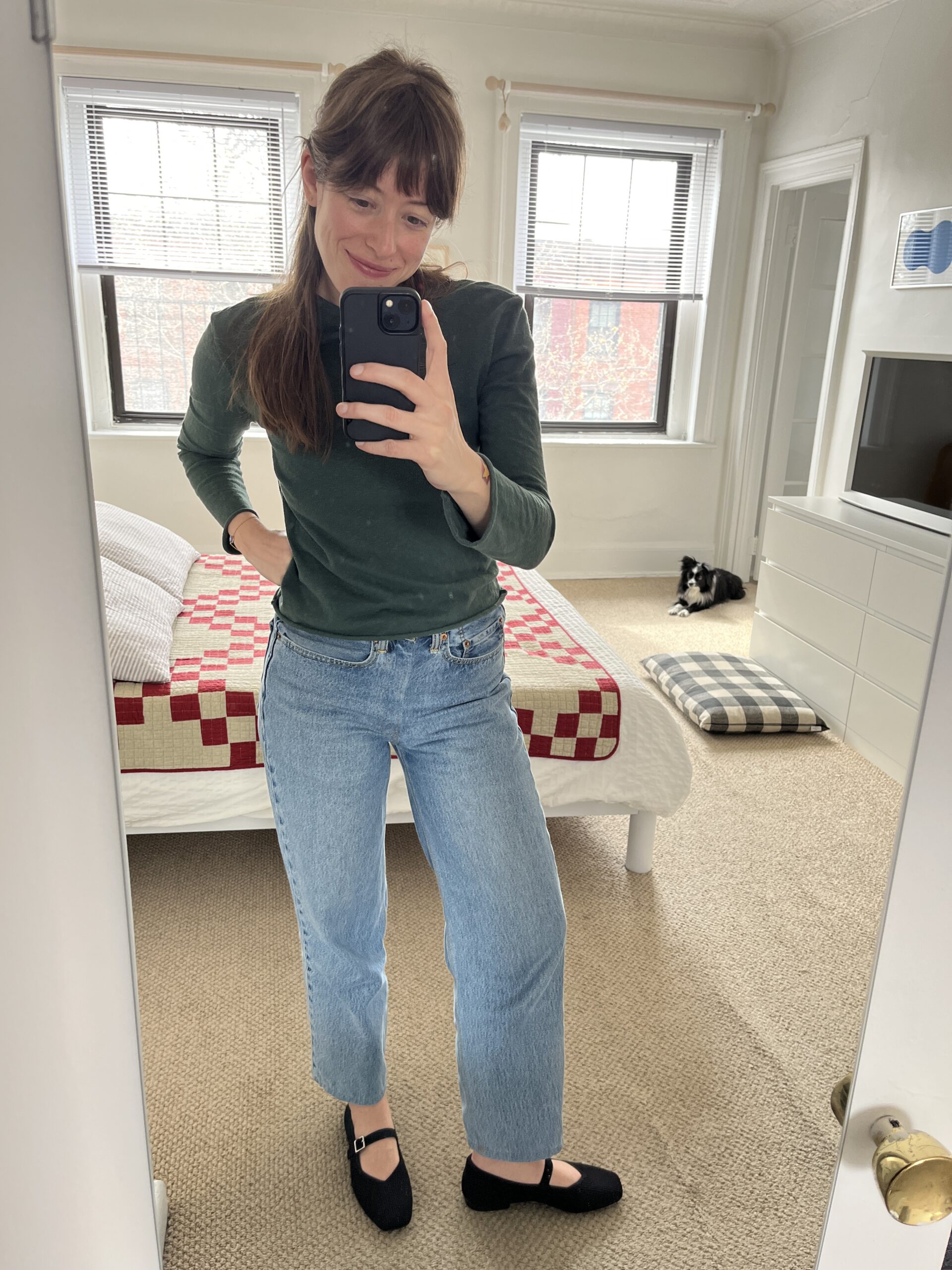 A woman taking a mirror selfie in a bedroom wearing a green top, light blue jeans, and black shoes. A dog lies on a checkered cushion in the background near the bed.