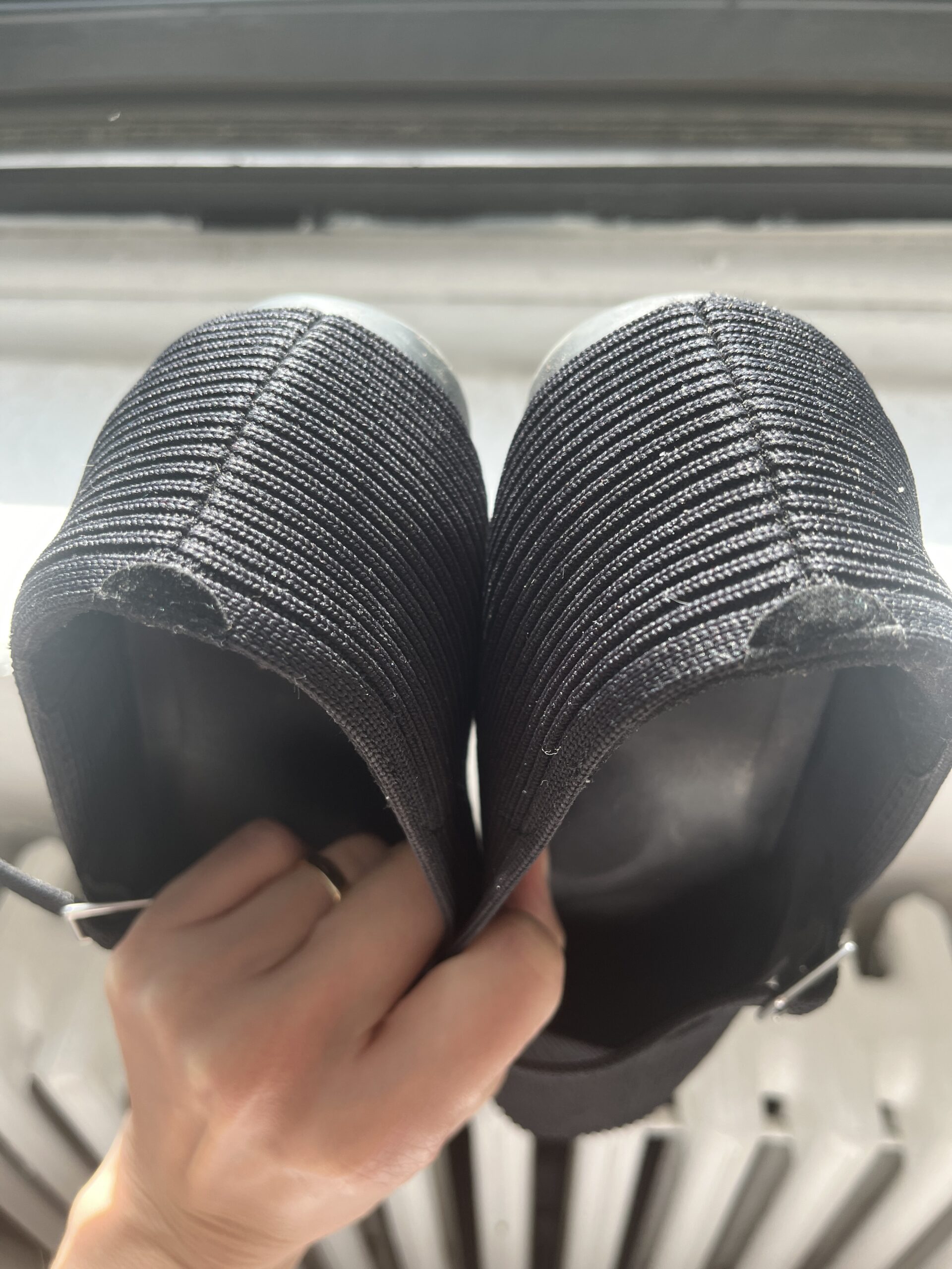 A person is holding a pair of black knit shoes by the heels, viewed from above. The shoes feature a ribbed texture.