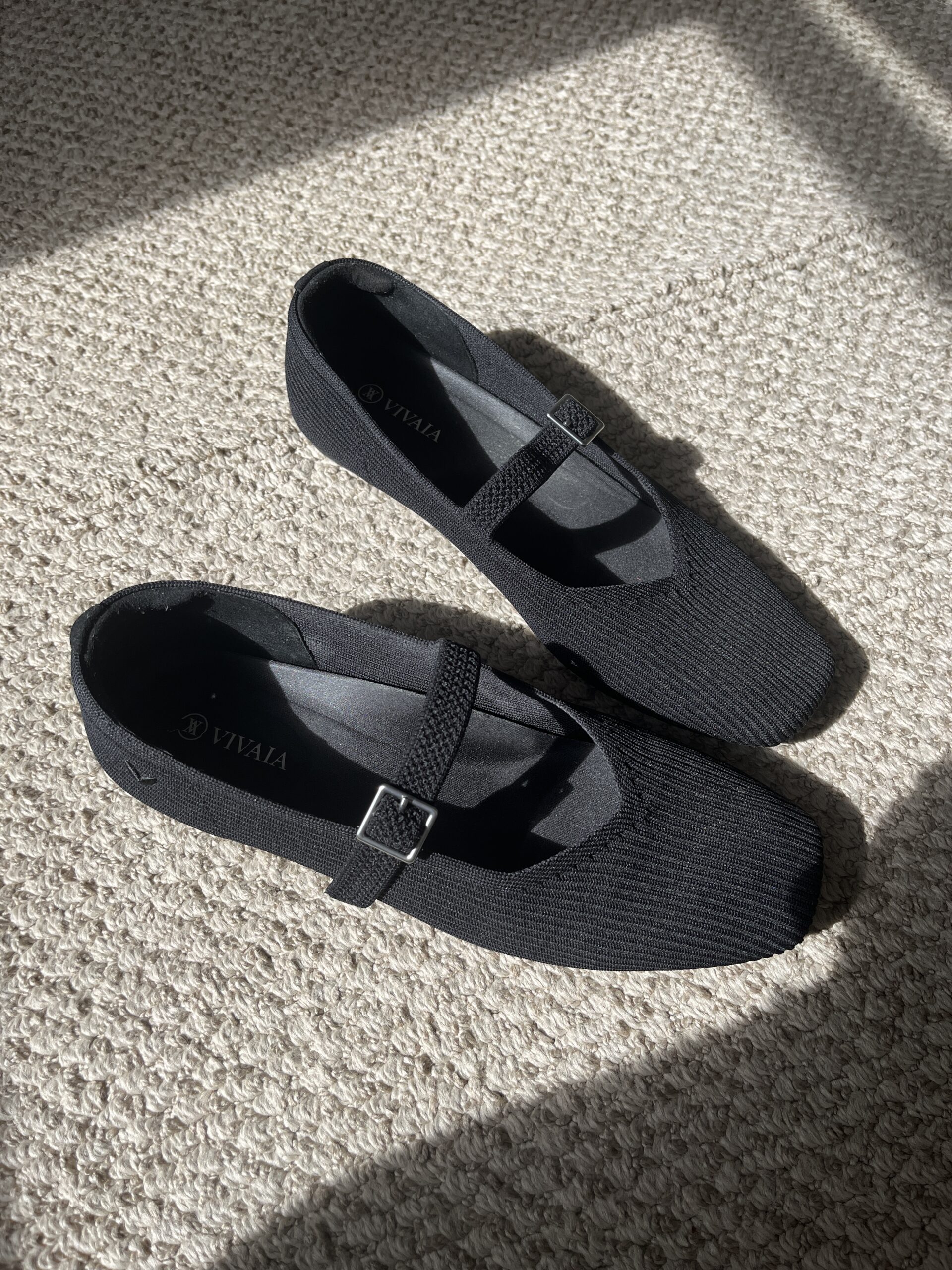 A pair of black Mary Jane shoes with a strap and buckle detail, placed on a beige textured carpet in a sunlit area.