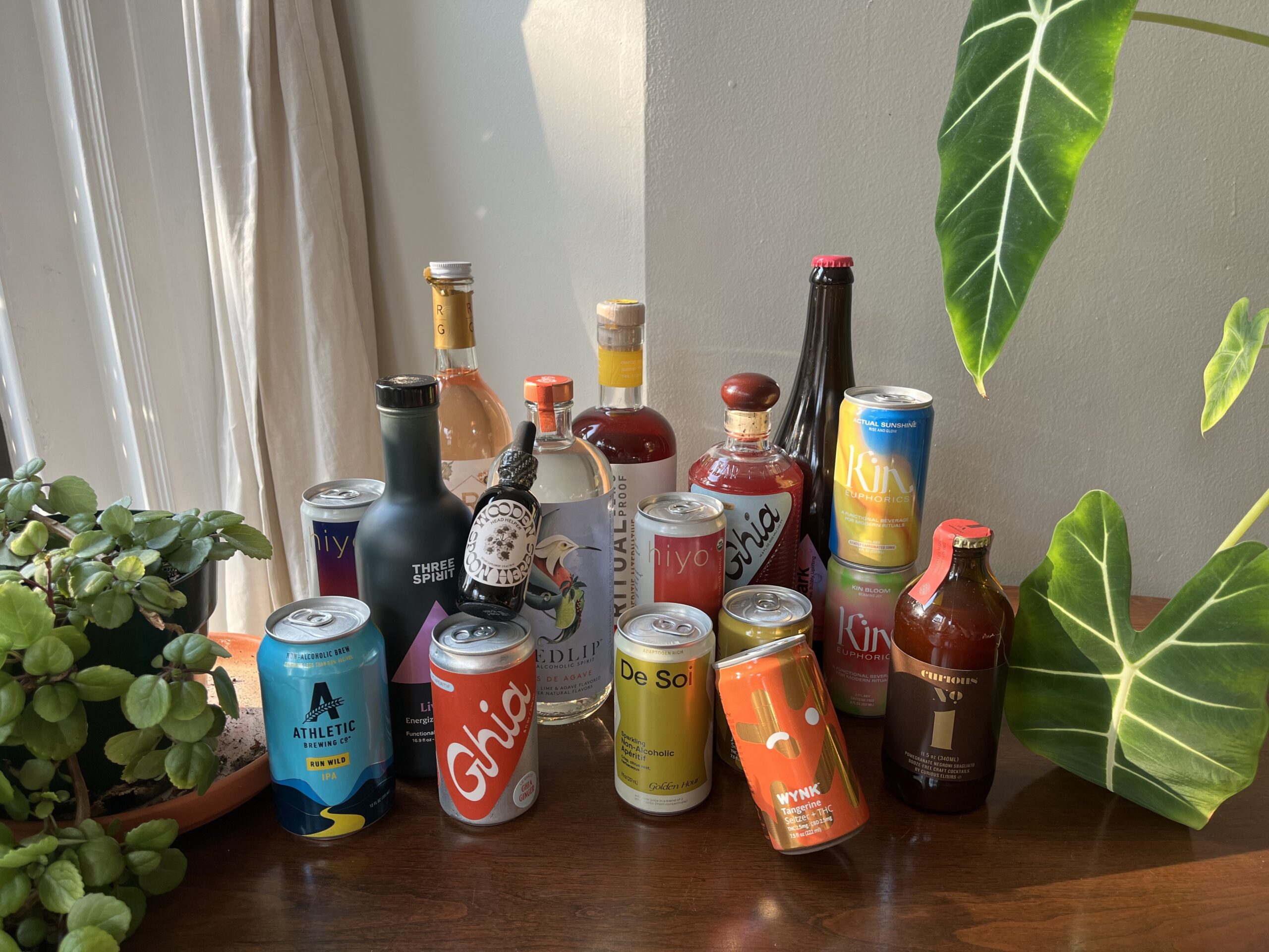 A variety of non-alcoholic beverages, including bottles and cans, displayed on a wooden surface near potted plants and a sunlit window.