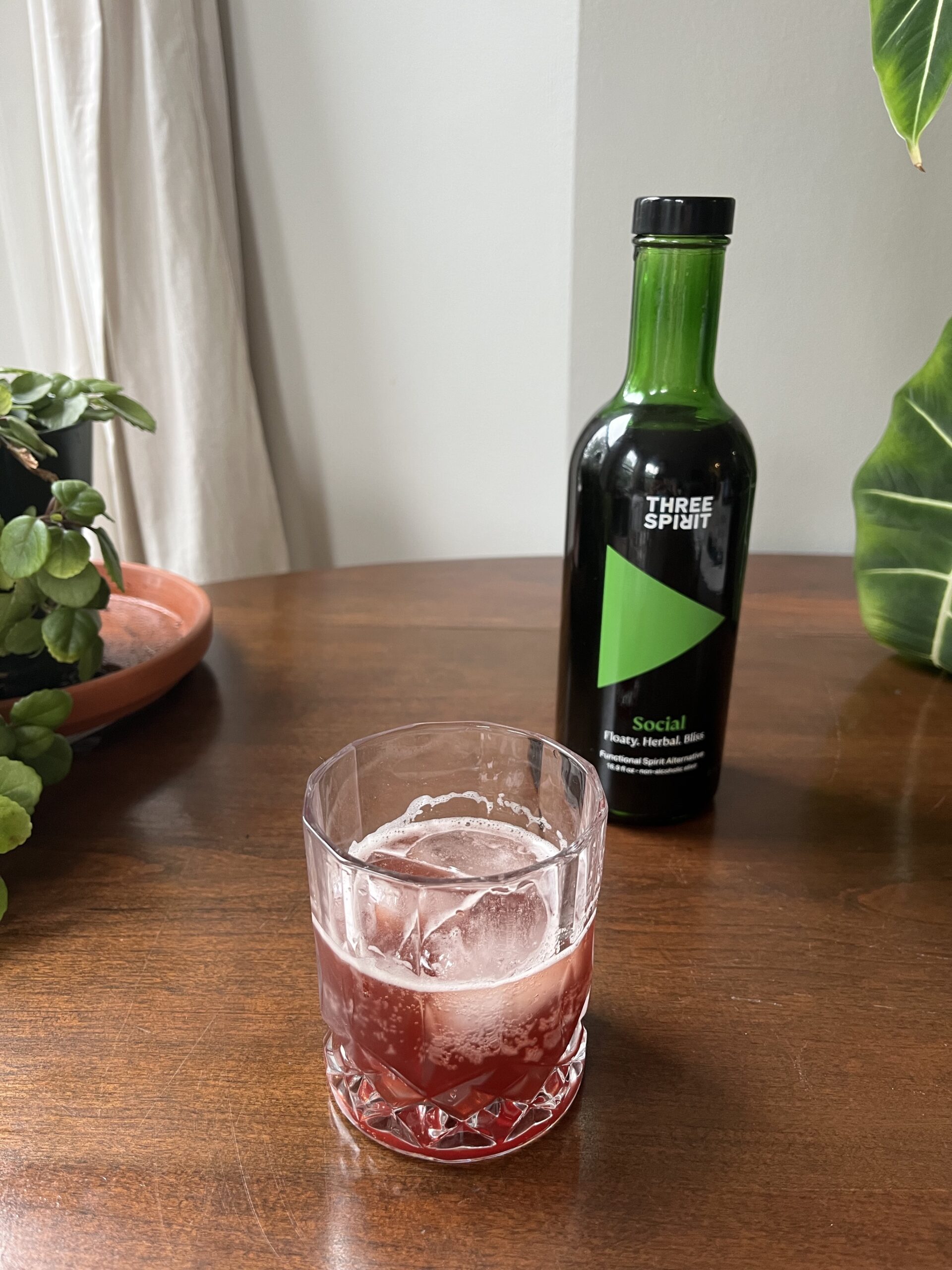 A bottle of Three Spirit Social Elixir stands on a wooden table beside a glass filled with a dark beverage and ice cubes, surrounded by green plants.