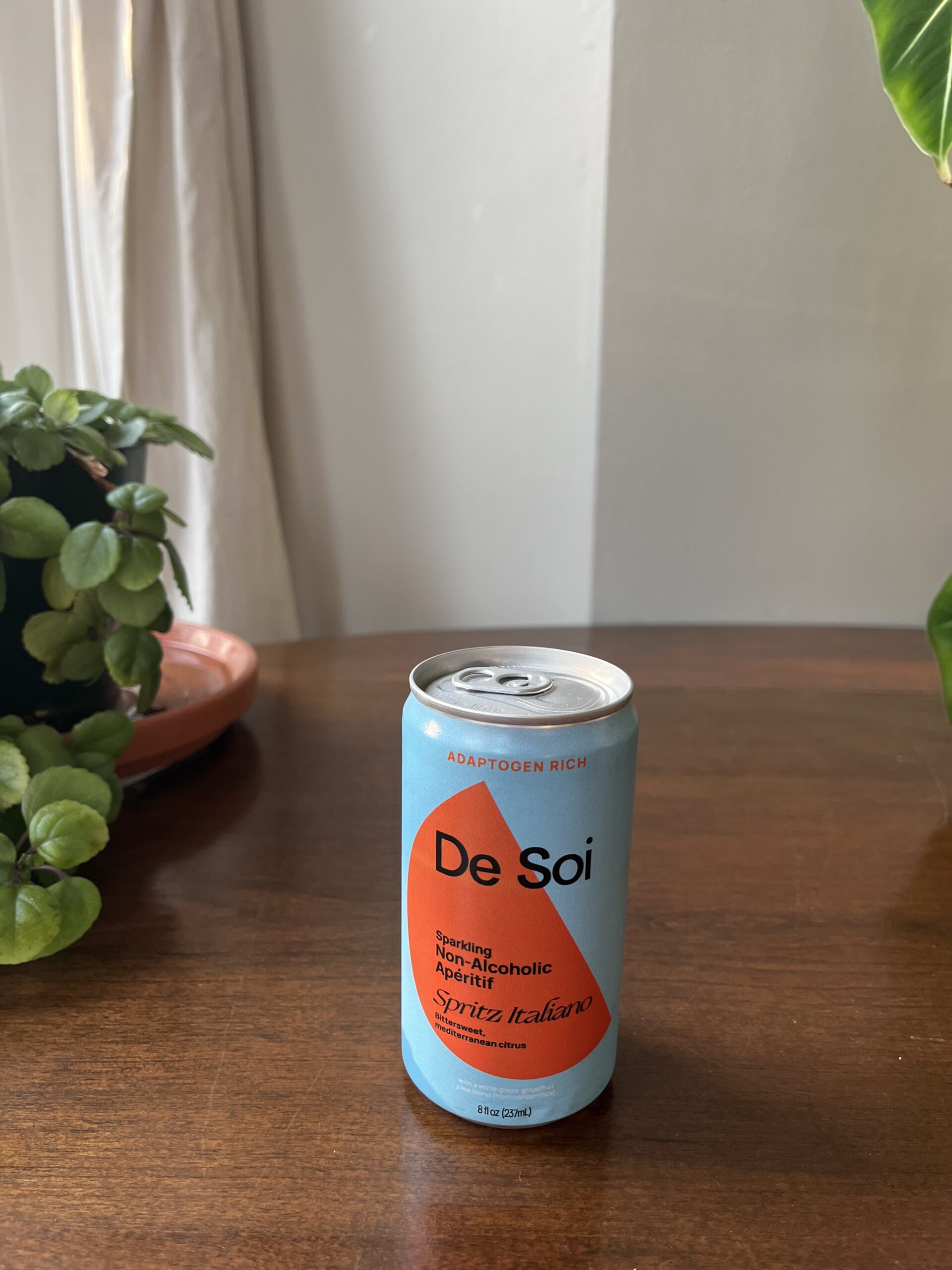 A can of De Soi Sparkling Non-Alcoholic Aperitif sits on a wooden table next to a plant.