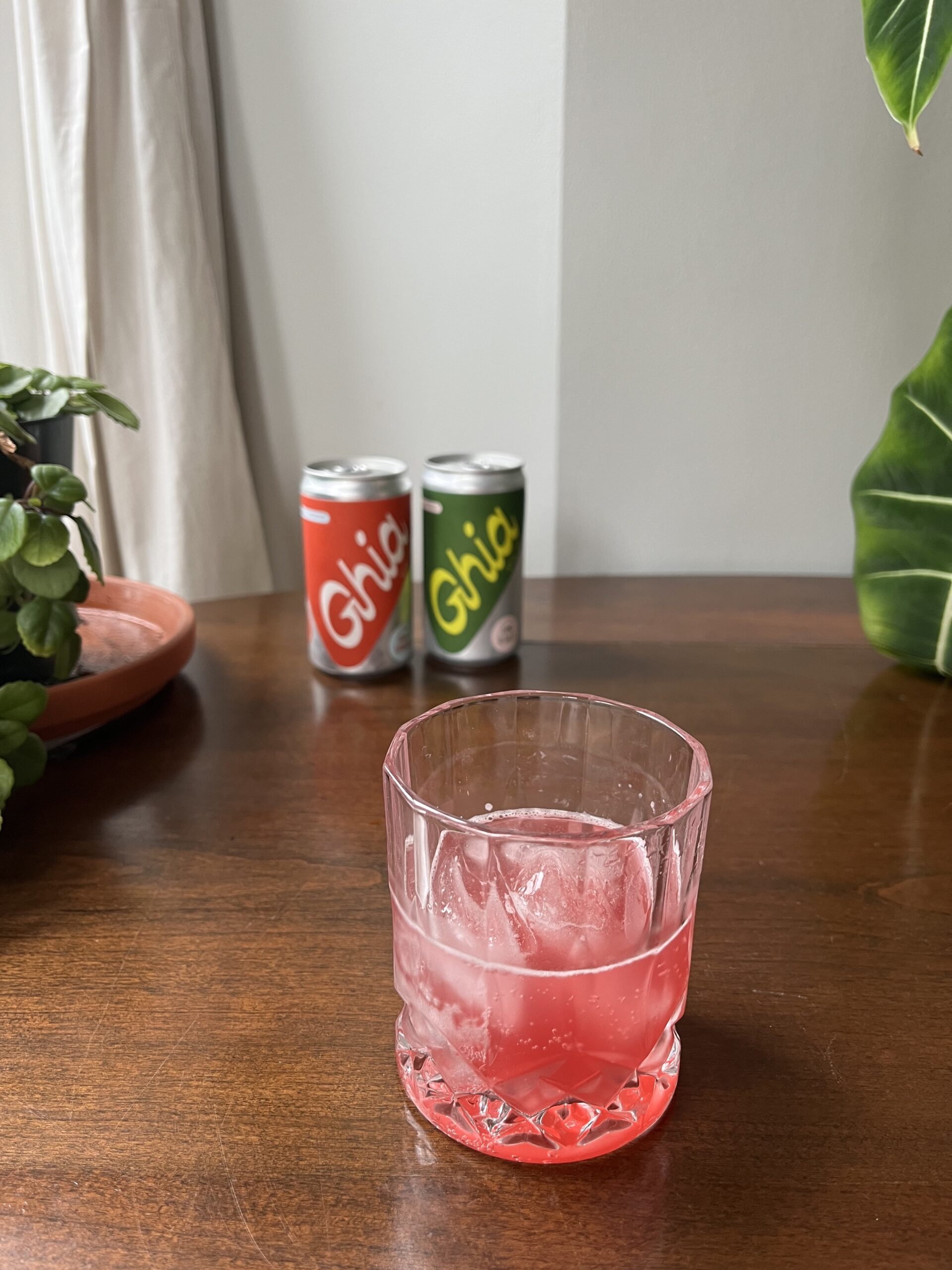 A glass of pink beverage with ice sits on a wooden table, with two cans of Ghia behind it. A potted plant is partially visible on the left.