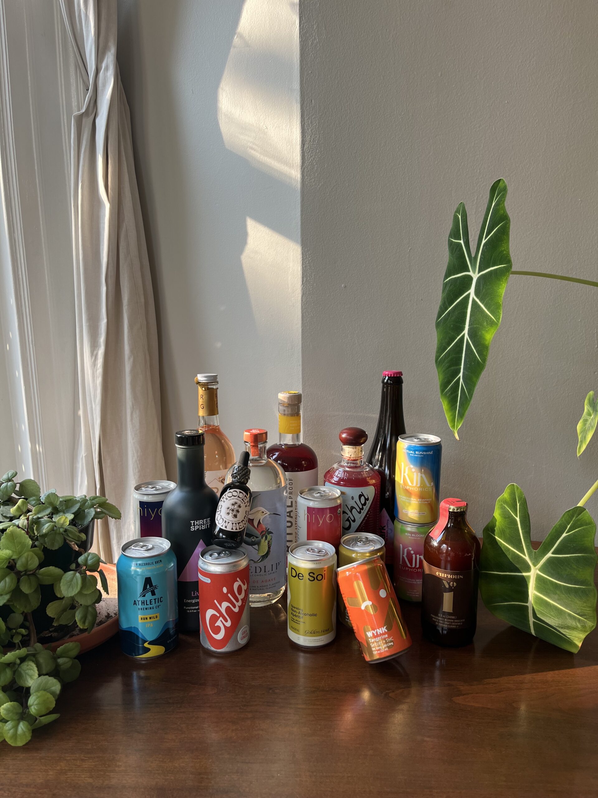 A collection of various beverages, including bottles and cans, displayed on a wooden surface with plants and a window in the background.