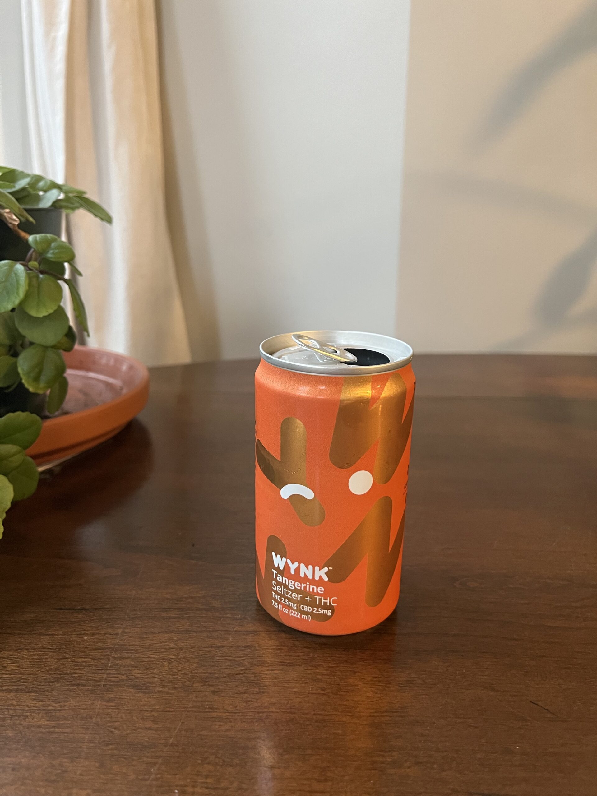 A tangerine-flavored Wynk cannabis seltzer can with an orange and white design is placed on a wooden table next to a potted plant.