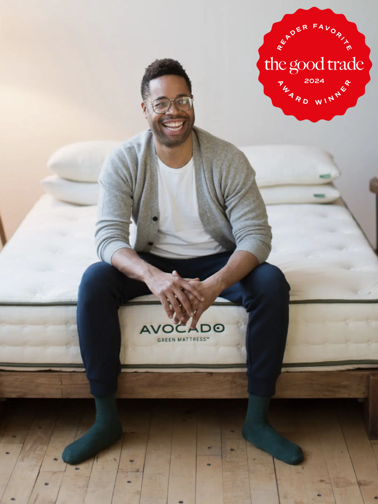 A person wearing a gray cardigan and dark pants sits on an Avocado Green Mattress in a wooden-framed bed, with a "The Good Trade 2024 Reader Favorite Award Winner" badge displayed.