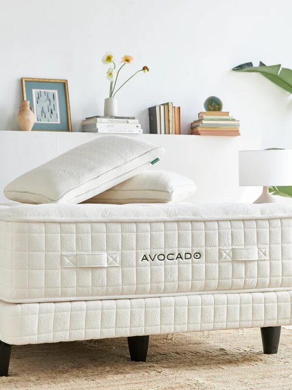 A neatly made bed with a cream-colored mattress labeled "AVOCADO," two pillows, a nightstand with a lamp, and shelves with books and decor in the background.