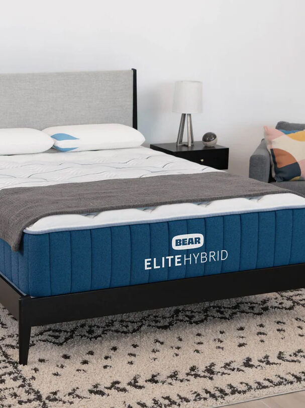 A neatly made bed with a blue mattress labeled "BEAR ELITE HYBRID" stands in a modern bedroom with a gray upholstered headboard, a nightstand, and a gray and white patterned rug underneath.