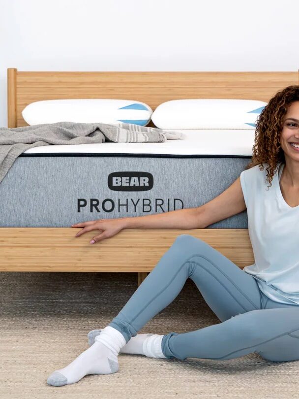 A woman in casual clothing sits on the floor next to a bed with a "Bear Pro Hybrid" mattress. She smiles and rests one hand on the mattress.