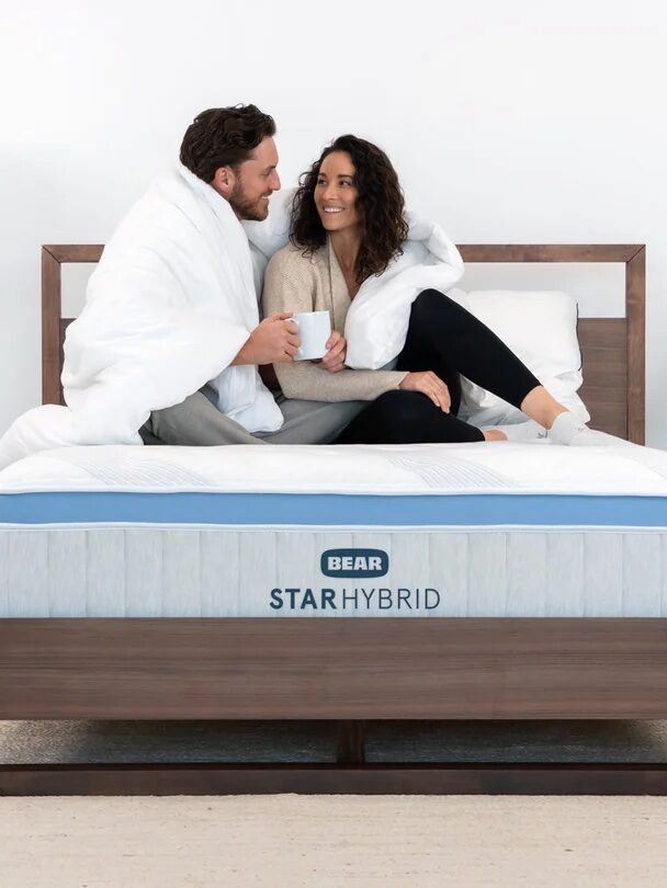 Two people sit on a bed wrapped in white blankets. They are smiling and holding mugs. The bed has a Bear Star Hybrid mattress. The room is minimally decorated with a lamp, plants, and a small nightstand.