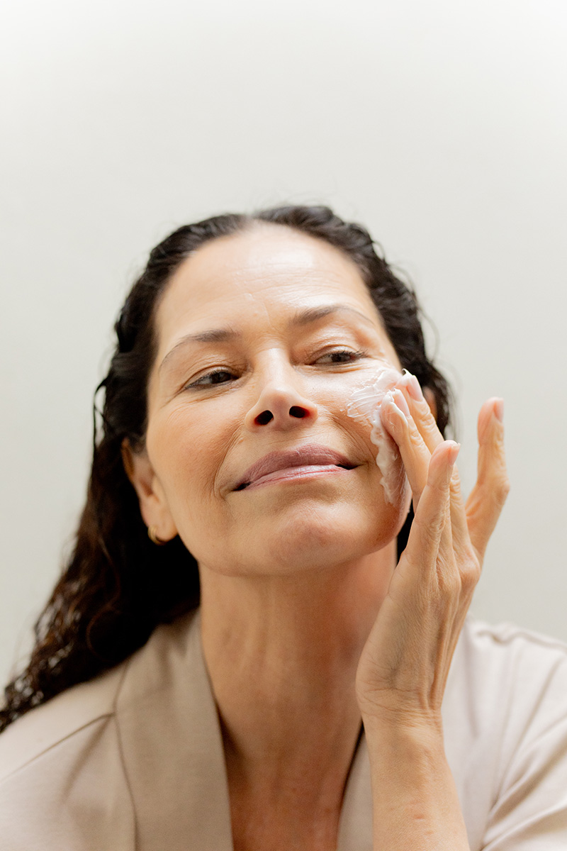 A woman applies a skincare product to her face with a gentle smile. She uses her right hand to spread the cream.