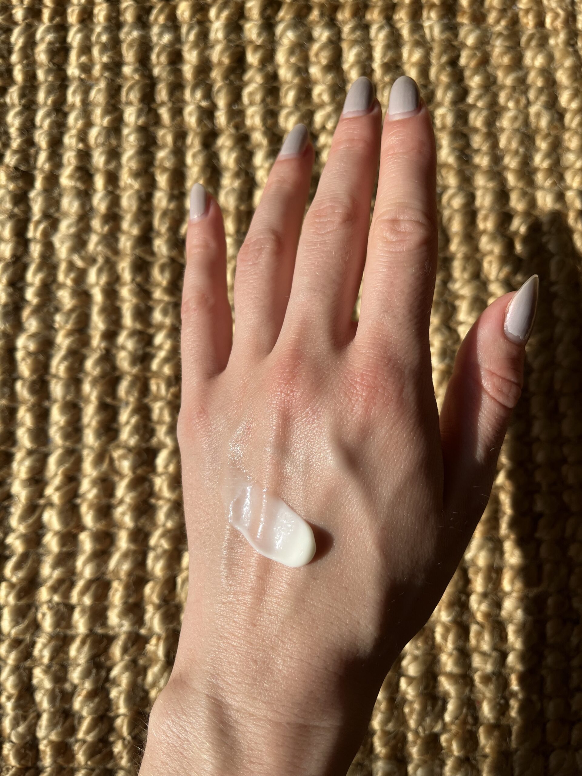 A hand with nude-colored nails displaying a dollop of white cream on the back, positioned against a woven, textured surface.