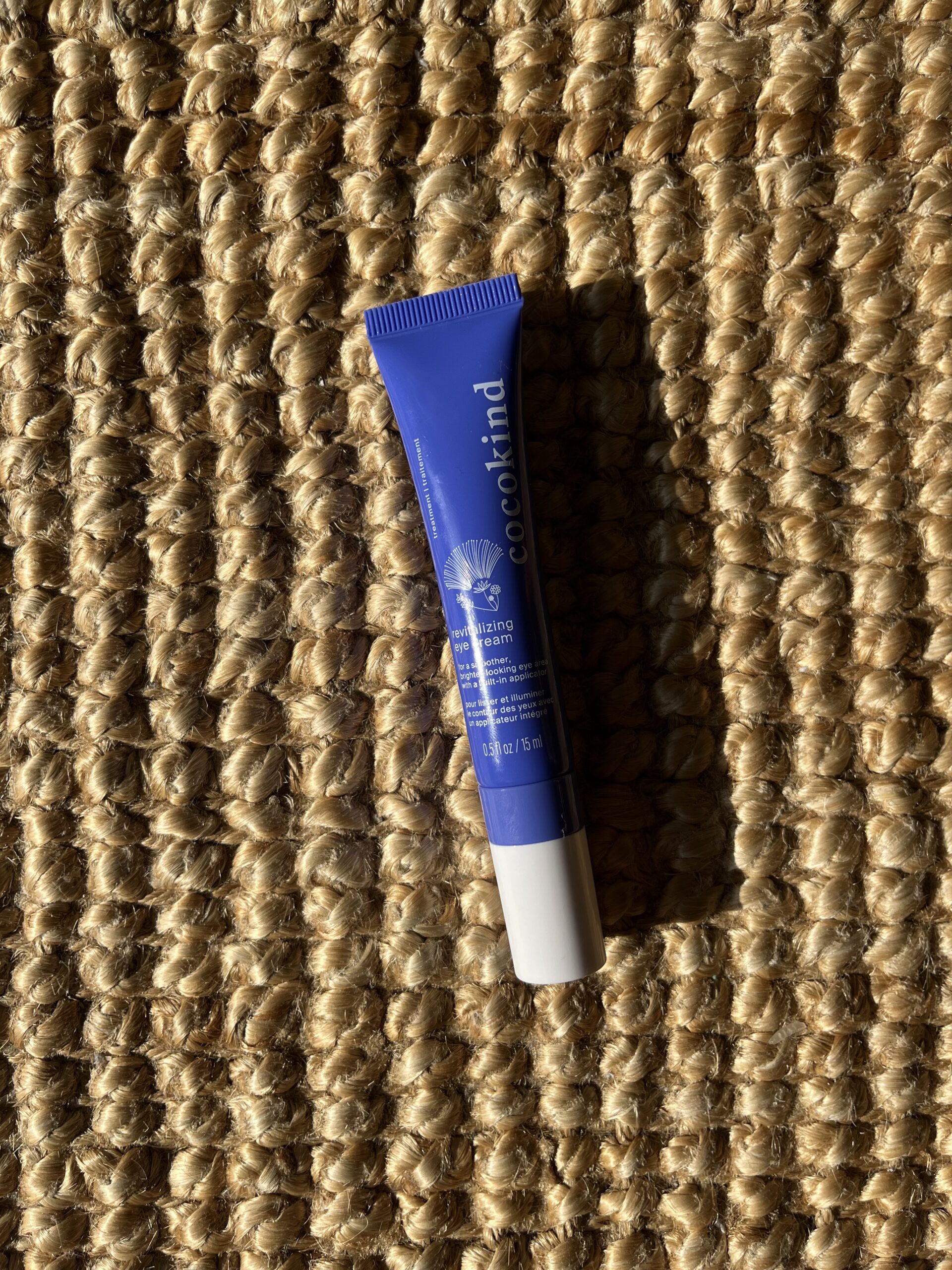 A blue tube of Cocokind skincare product lies on a woven beige mat, with text and logo visible on the tube.