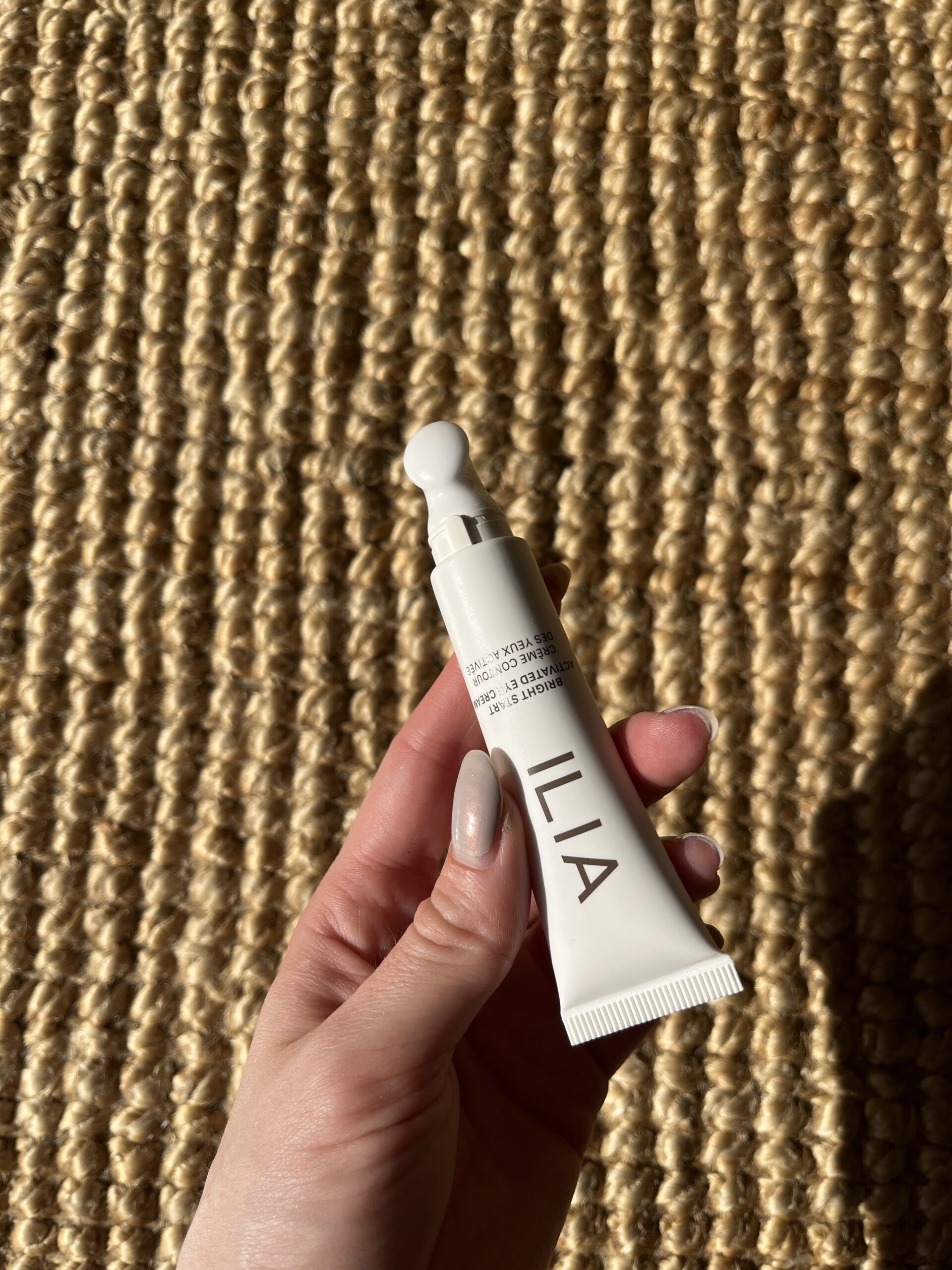 A hand holds a white tube labeled "ILIA" against a woven fabric background.