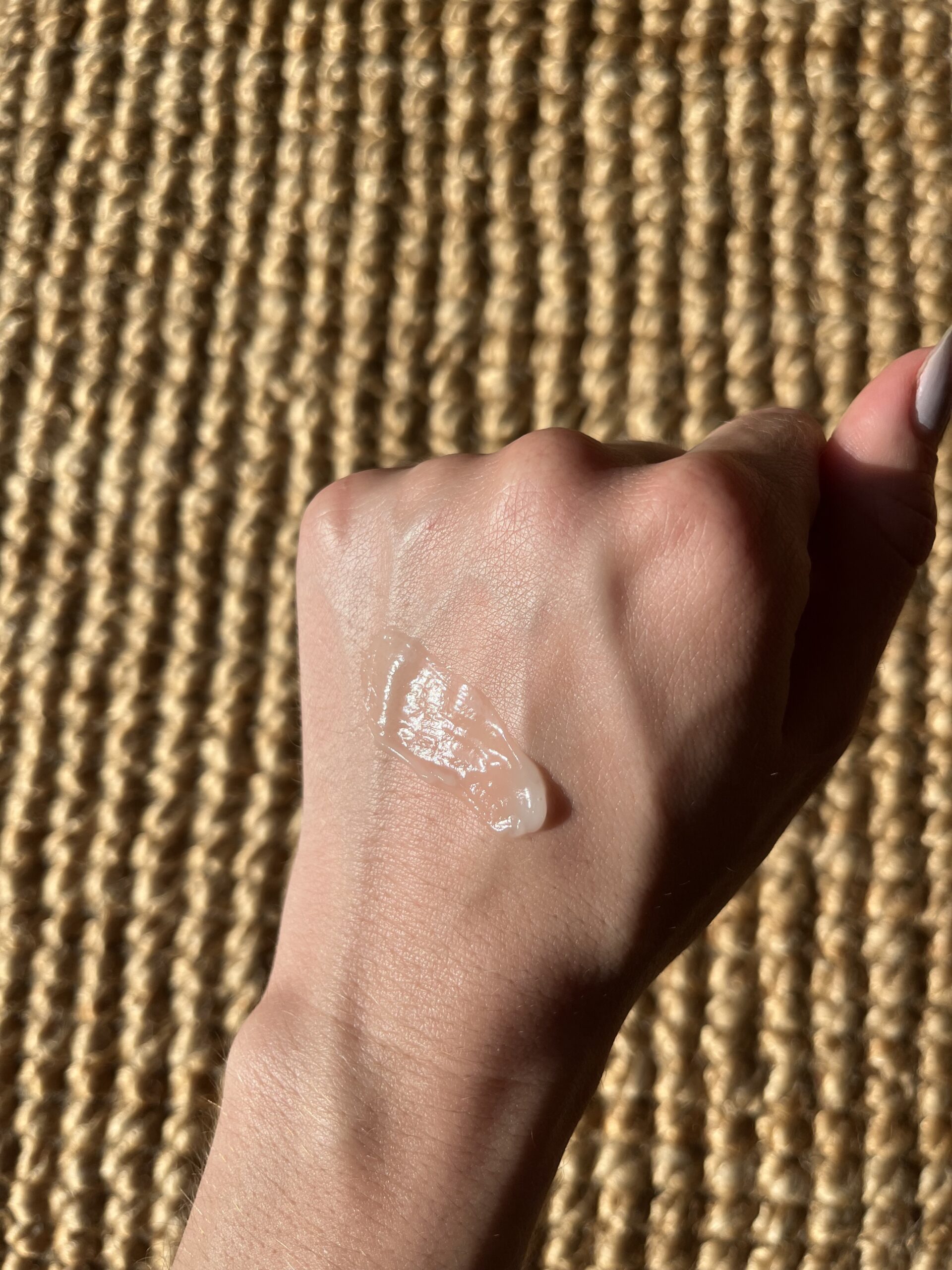 A close-up image of a person's hand with a dollop of clear gel on the back, resting on a textured, woven surface.