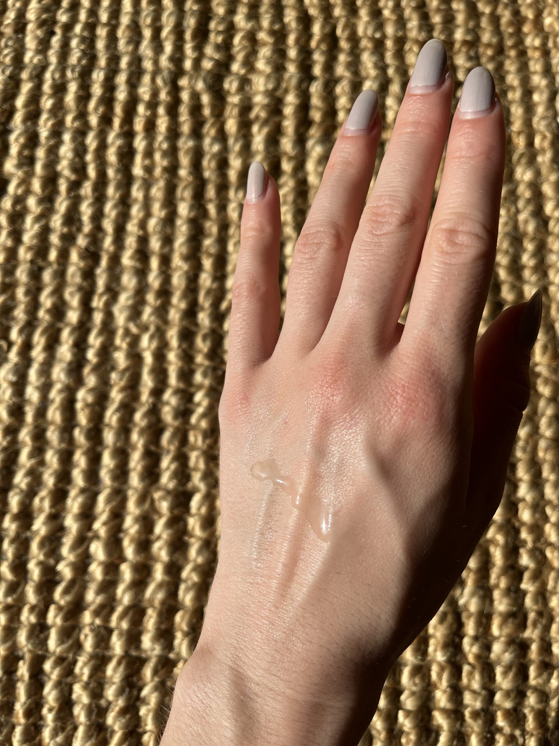 A hand with gray-painted nails is shown with a small amount of clear liquid on its back, resting against a textured, woven surface illuminated by sunlight.