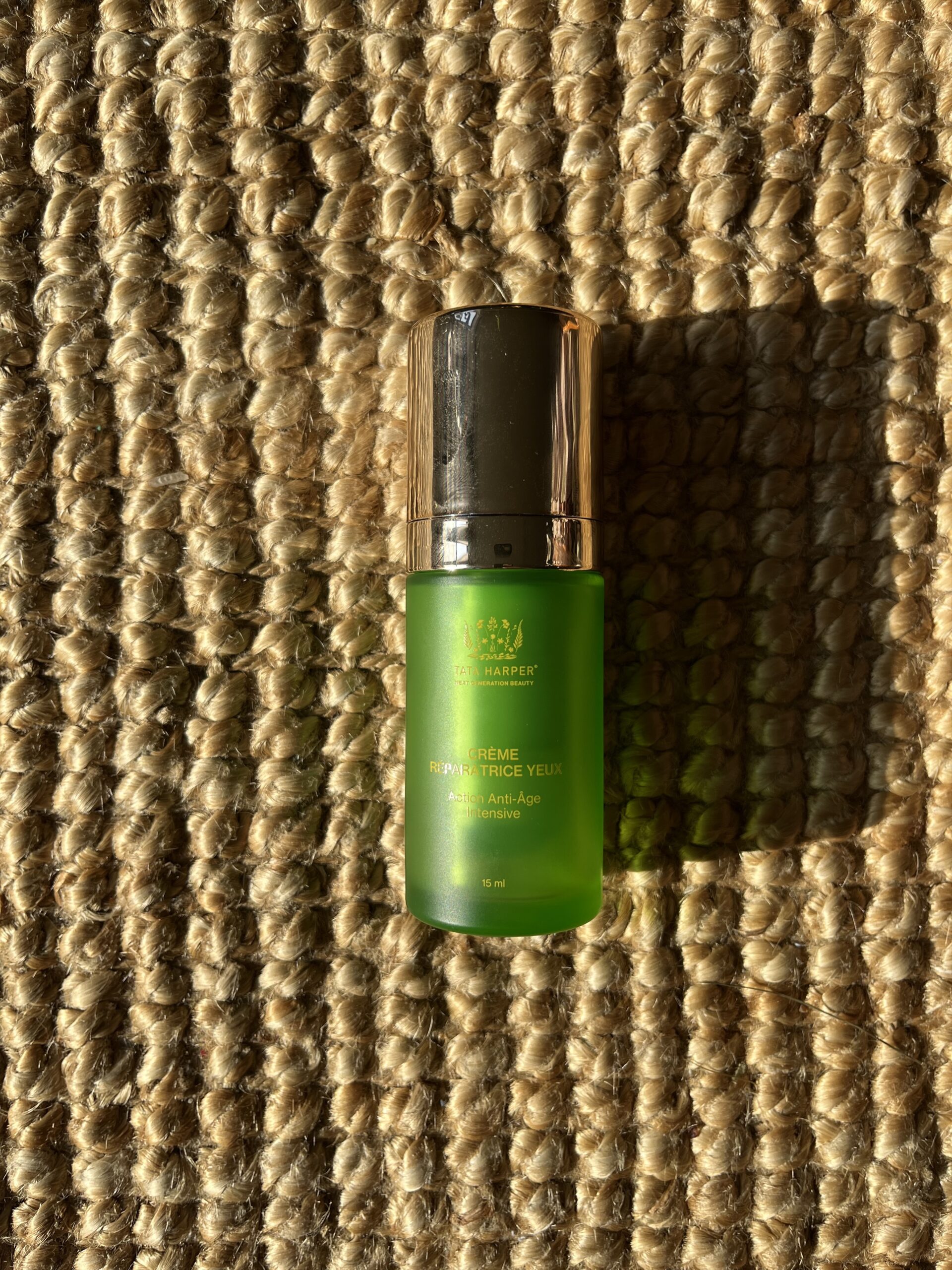 A green cylindrical bottle of Tata Harper eye cream with a silver cap is placed on a beige woven surface, illuminated by sunlight.