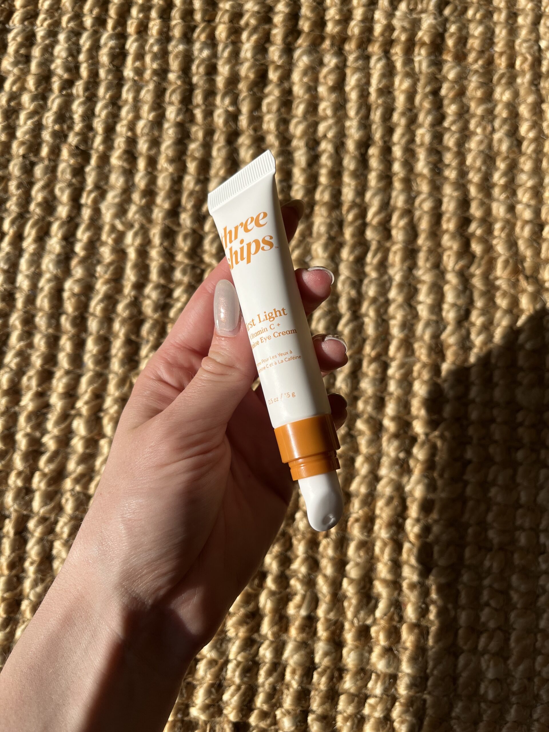 Hand holding a white tube of skincare product labeled "Three Ships" against a woven textured background.