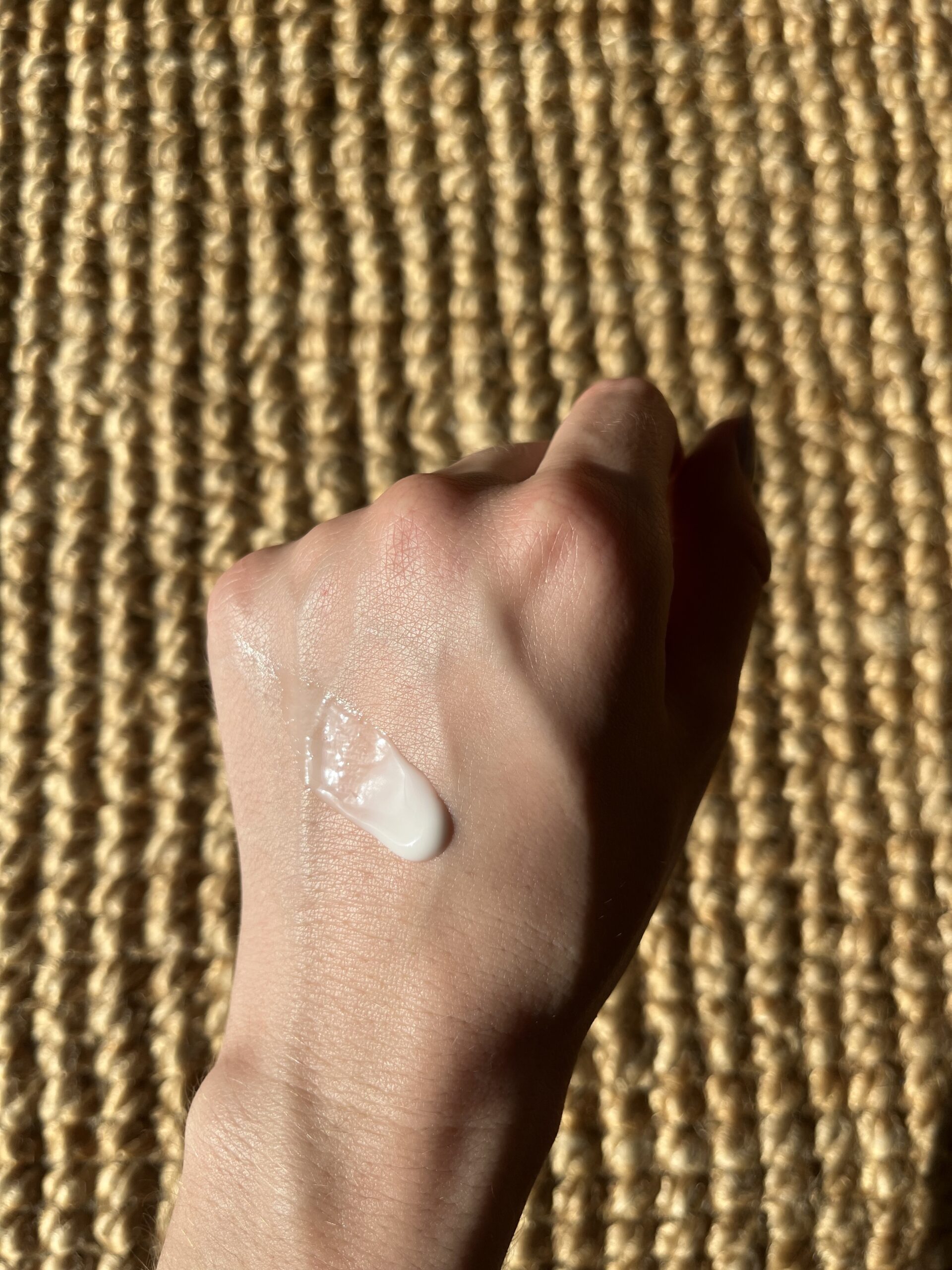 A hand with a small amount of white lotion applied on the back, resting on a woven beige mat.