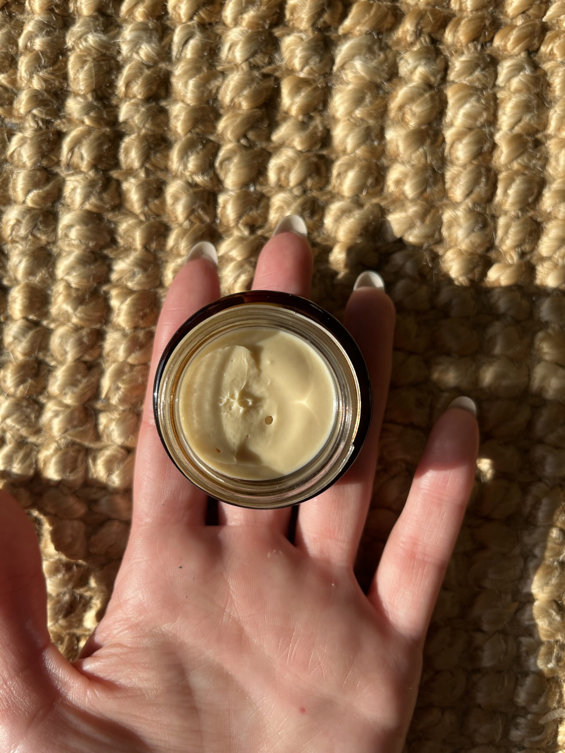 A hand with manicured nails holds an open container of a beige cream against a textured, woven surface.