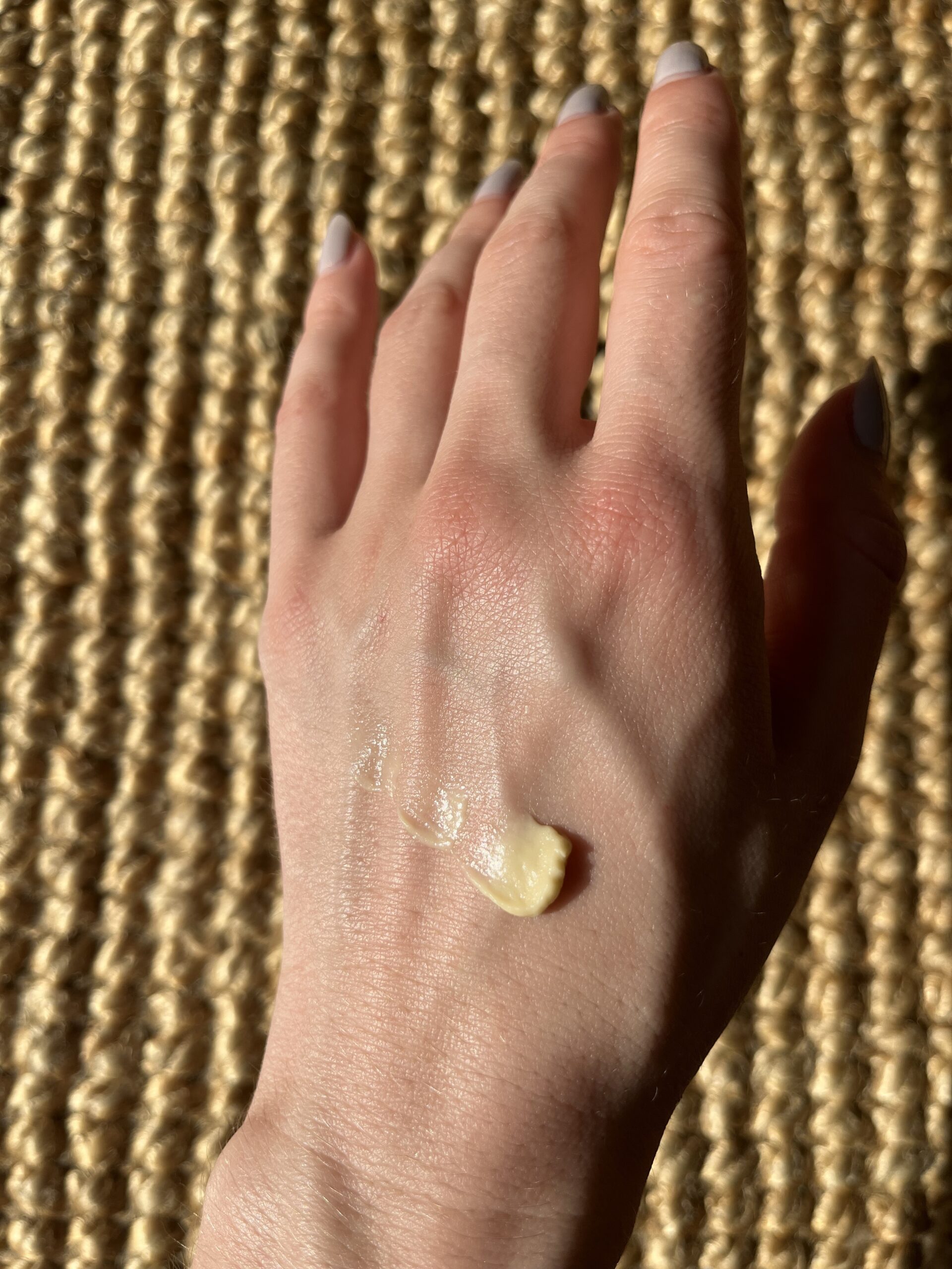 A close-up of a hand with a small amount of cream applied on the back, resting on a woven textured surface.