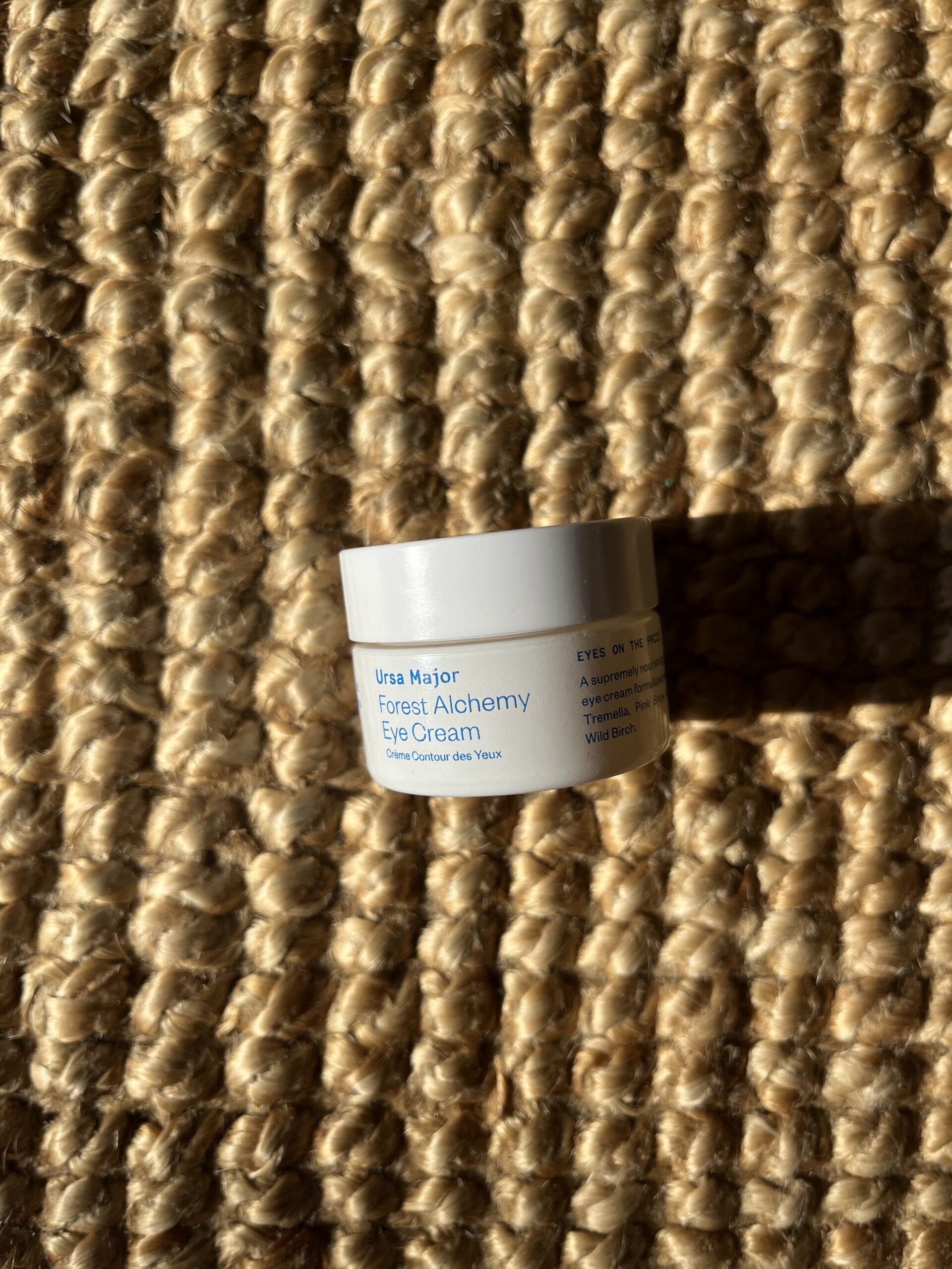 A small container of Ursa Major Forest Alchemy Eye Cream sits on a woven, beige textured surface, illuminated by sunlight.