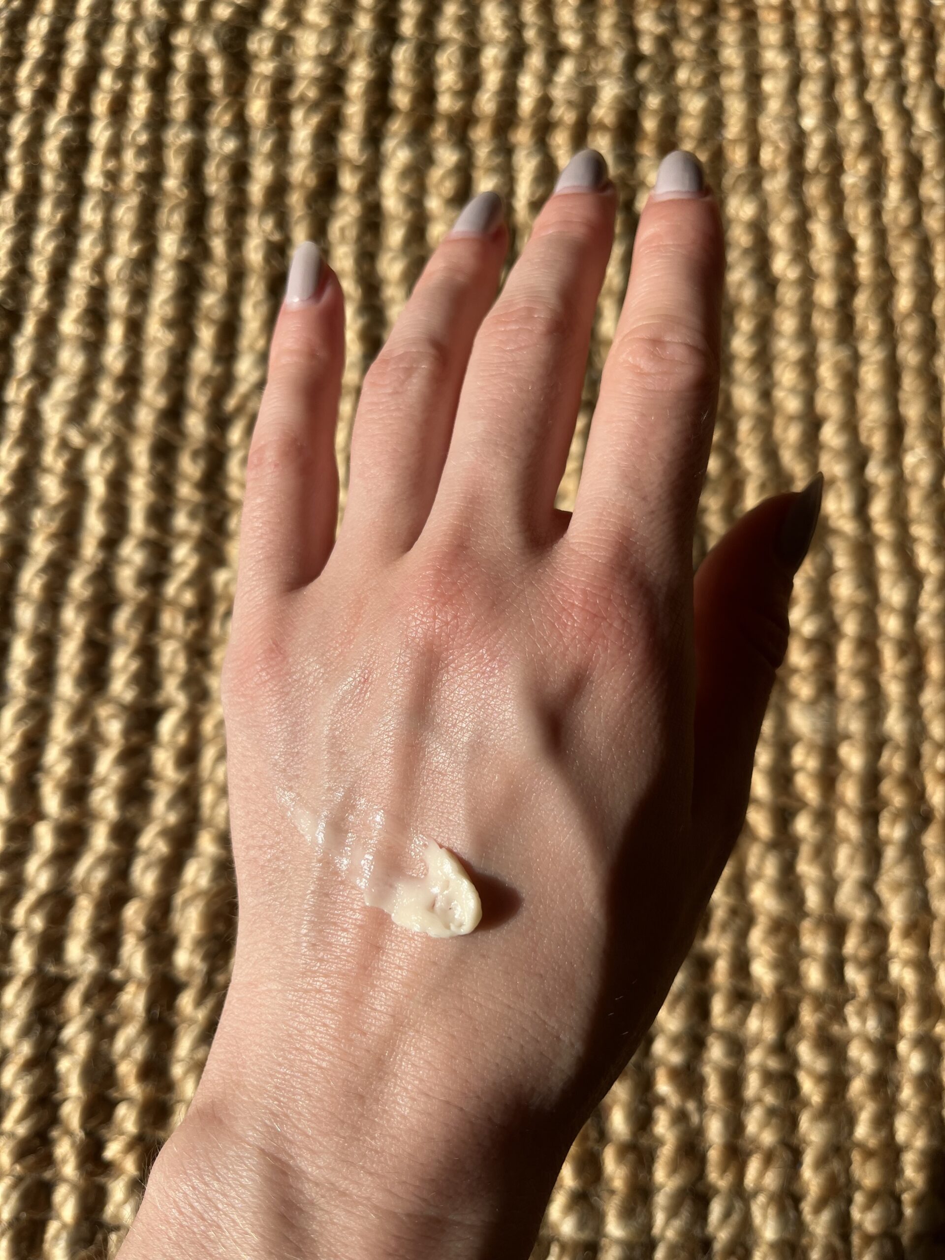 A hand with light skin tone, topped with a small dollop of white lotion, is resting on a woven surface.