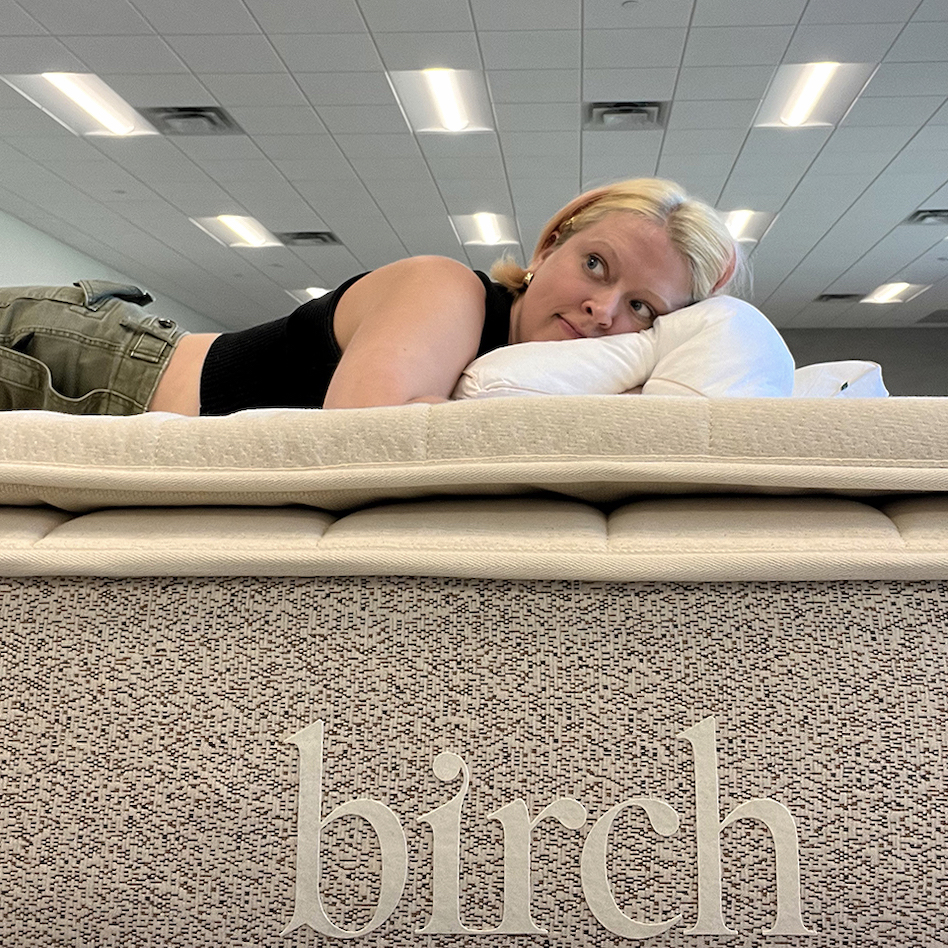 A person with short blonde hair, wearing a black top and green pants, lies on a mattress labeled "birch" in a spacious room with ceiling lights.