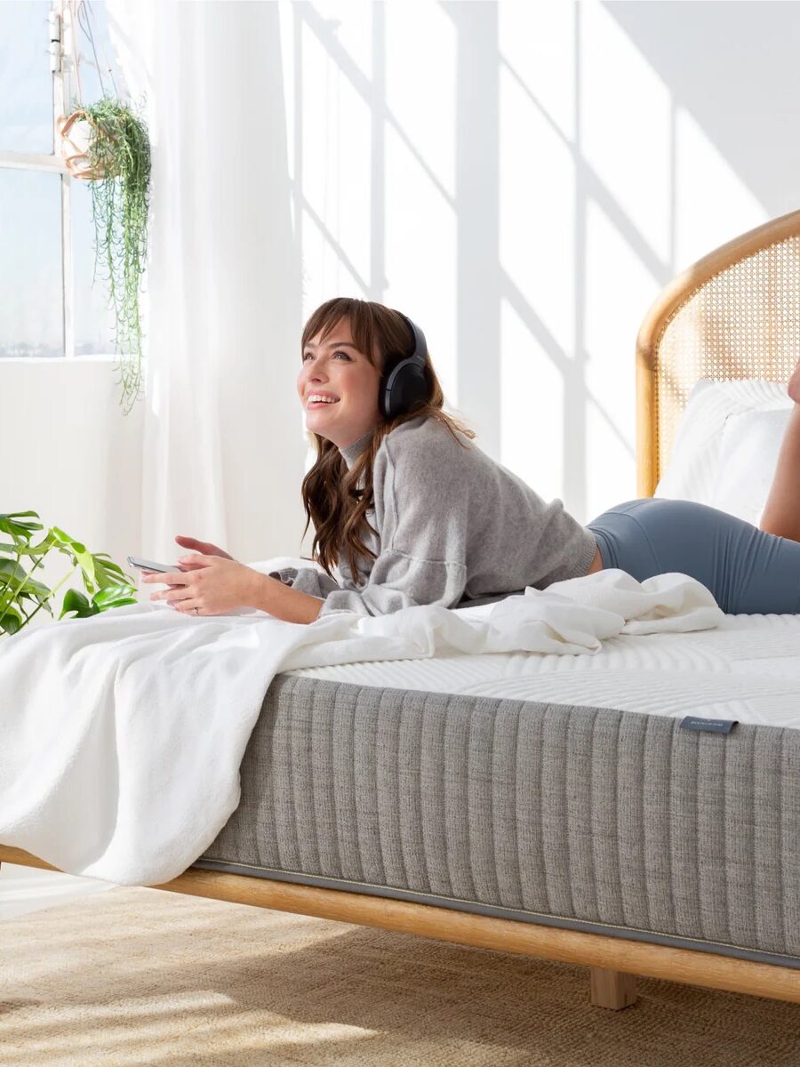 A person lying on a bed with headphones, holding a game controller, surrounded by indoor plants in a well-lit room.