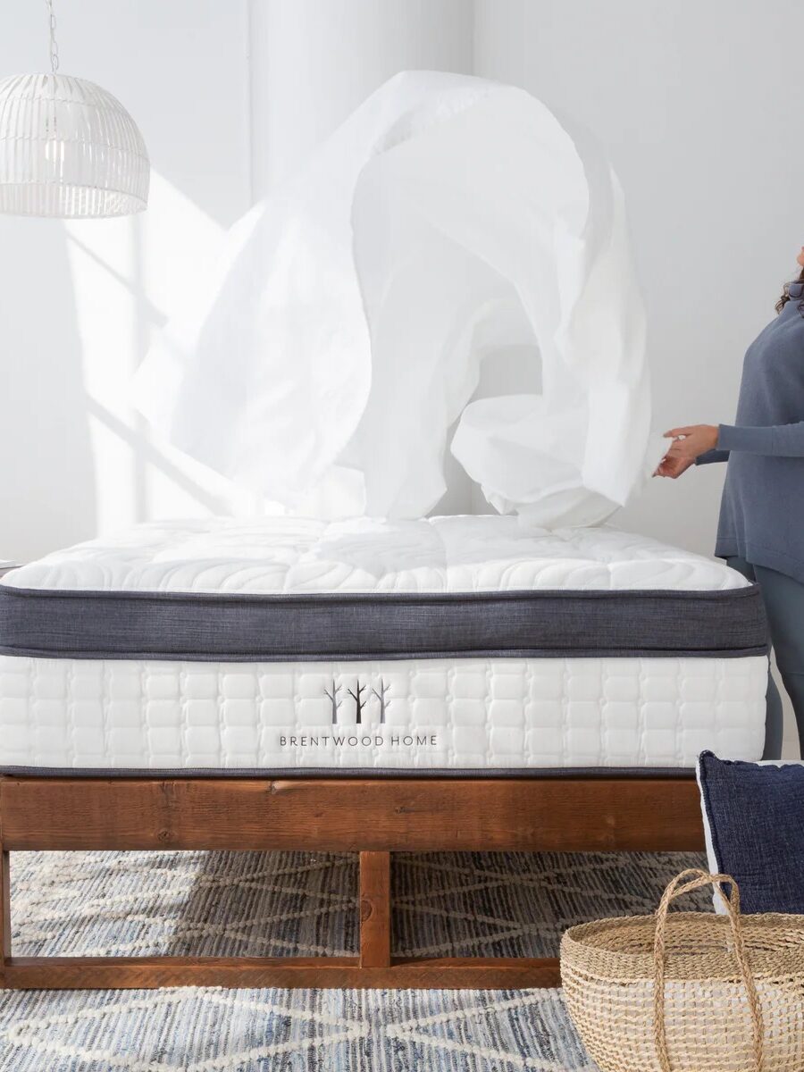 A woman makes a bed, tossing a white sheet over a mattress labeled "Brentwood Home" in a bright, well-lit room with wooden furniture and a patterned rug.