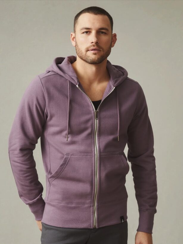 A man in a purple hoodie standing against a neutral background, facing the camera with a serious expression.