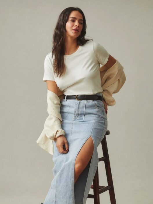 A model leaning against a stool wearing a white t-shirt and a denim maxi skirt with a slit from American Giant. 