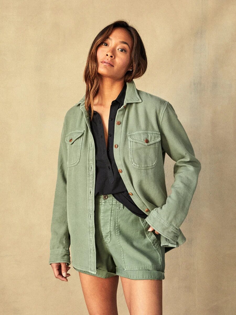 A woman wearing a green jacket over a black top, standing against a beige background, looking at the camera with a slight tilt of her head.