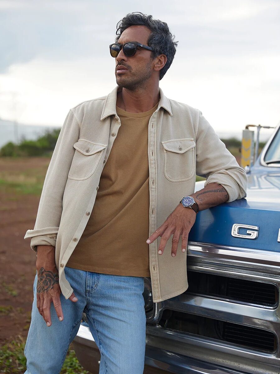 Man in sunglasses and casual outfit leaning on a vintage car in an open field.