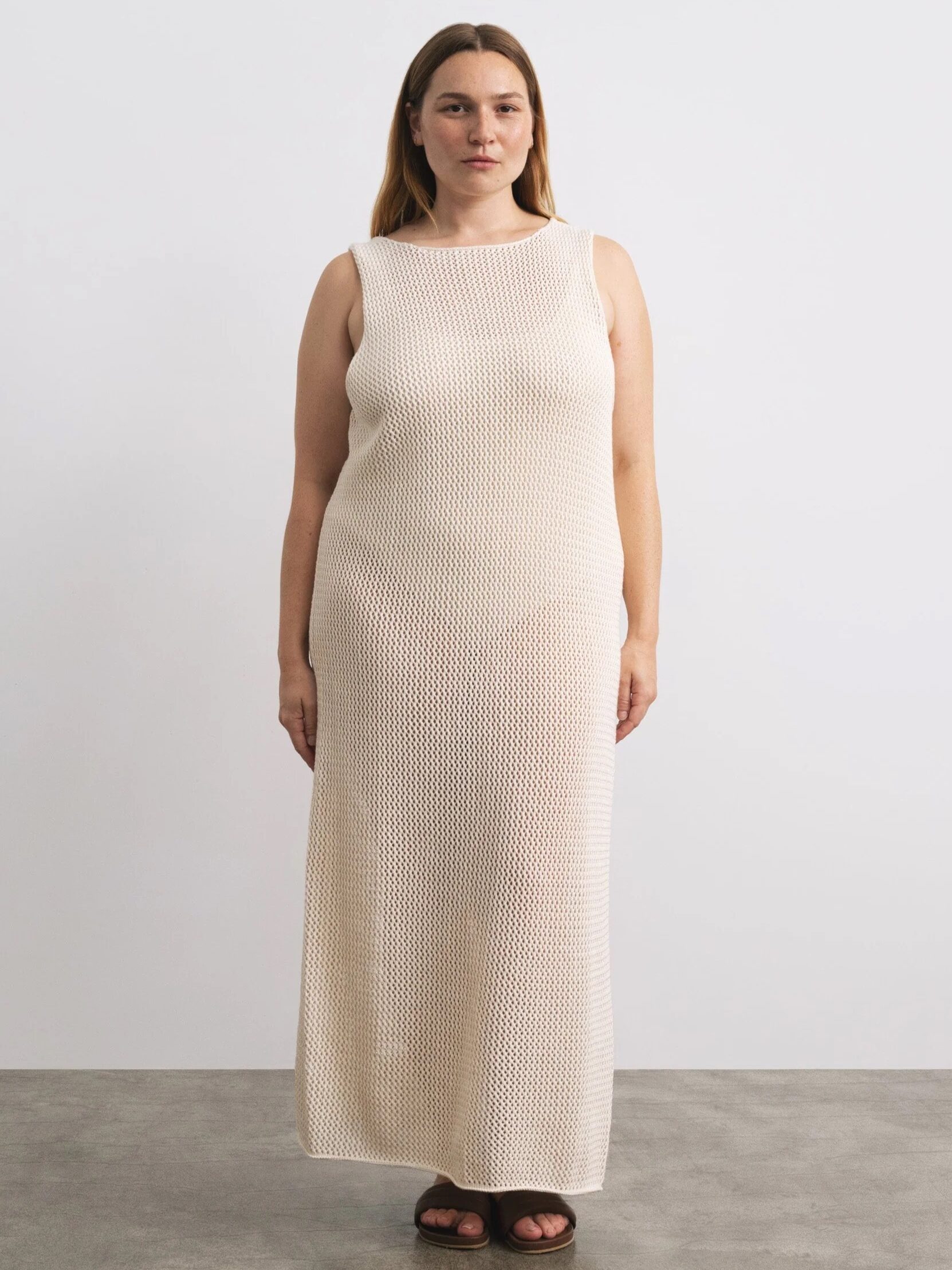 A woman in a beige sleeveless maxi dress stands against a plain background, looking at the camera with a neutral expression.