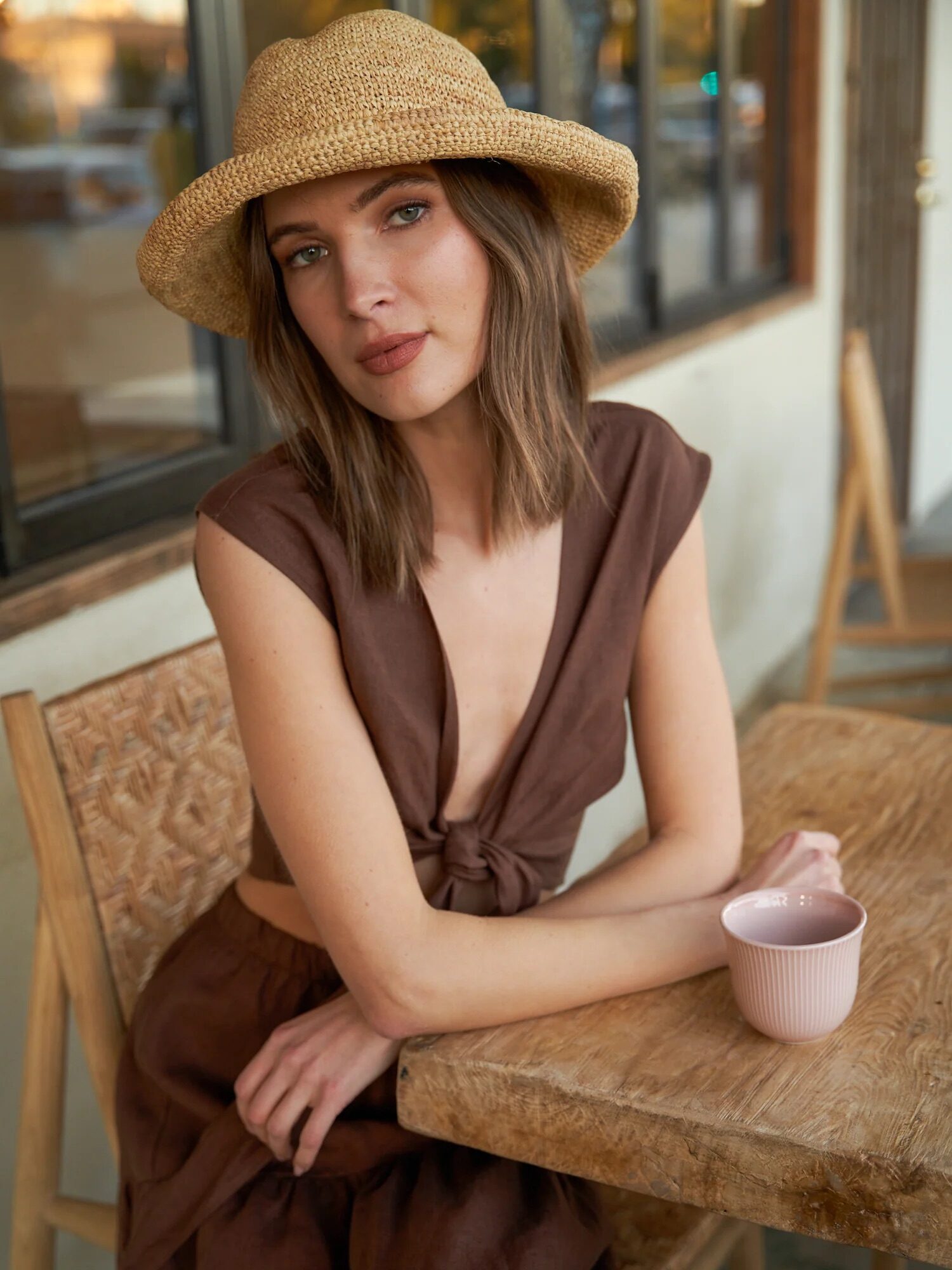 A woman in a straw hat and brown dress sitting at a cafe table holding a pink cup.