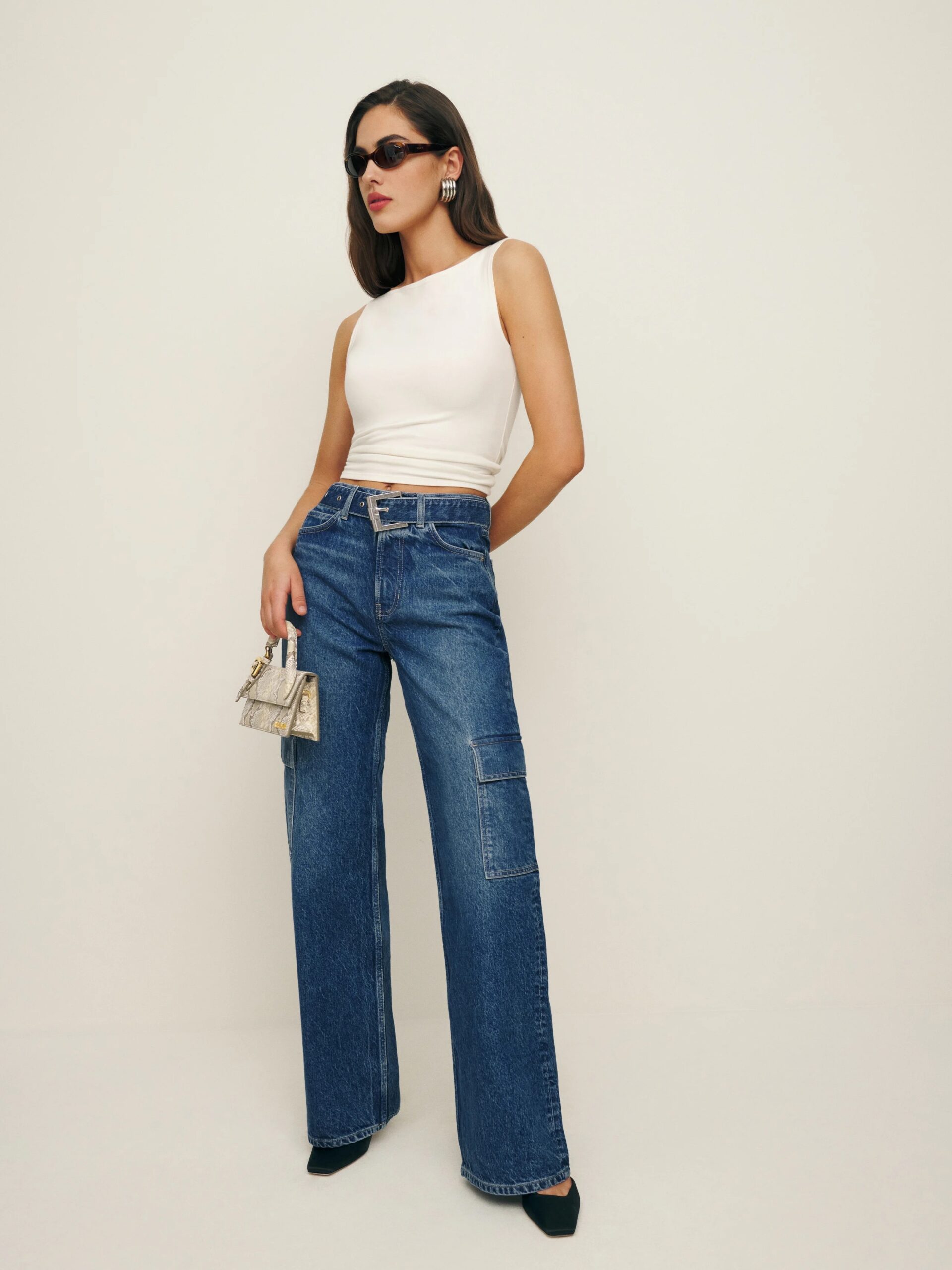 A woman stands against a plain background wearing a white sleeveless top, high-waisted blue jeans, black heels, sunglasses, and holding a small silver handbag. She has long hair and is posing confidently.