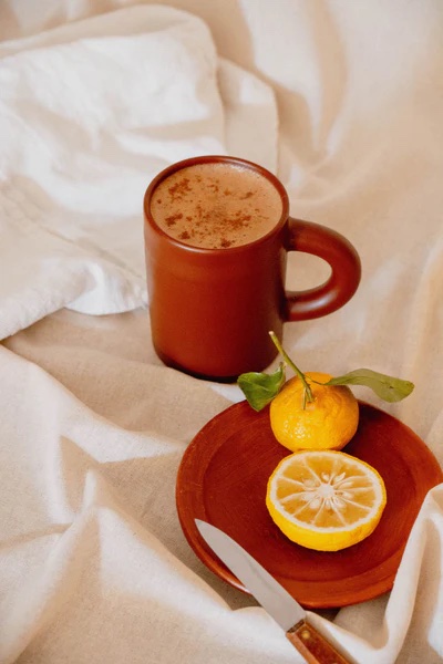 A brown mug with a frothy drink, a small orange and a lemon slice on a wooden plate with a knife, all placed on a light fabric surface.