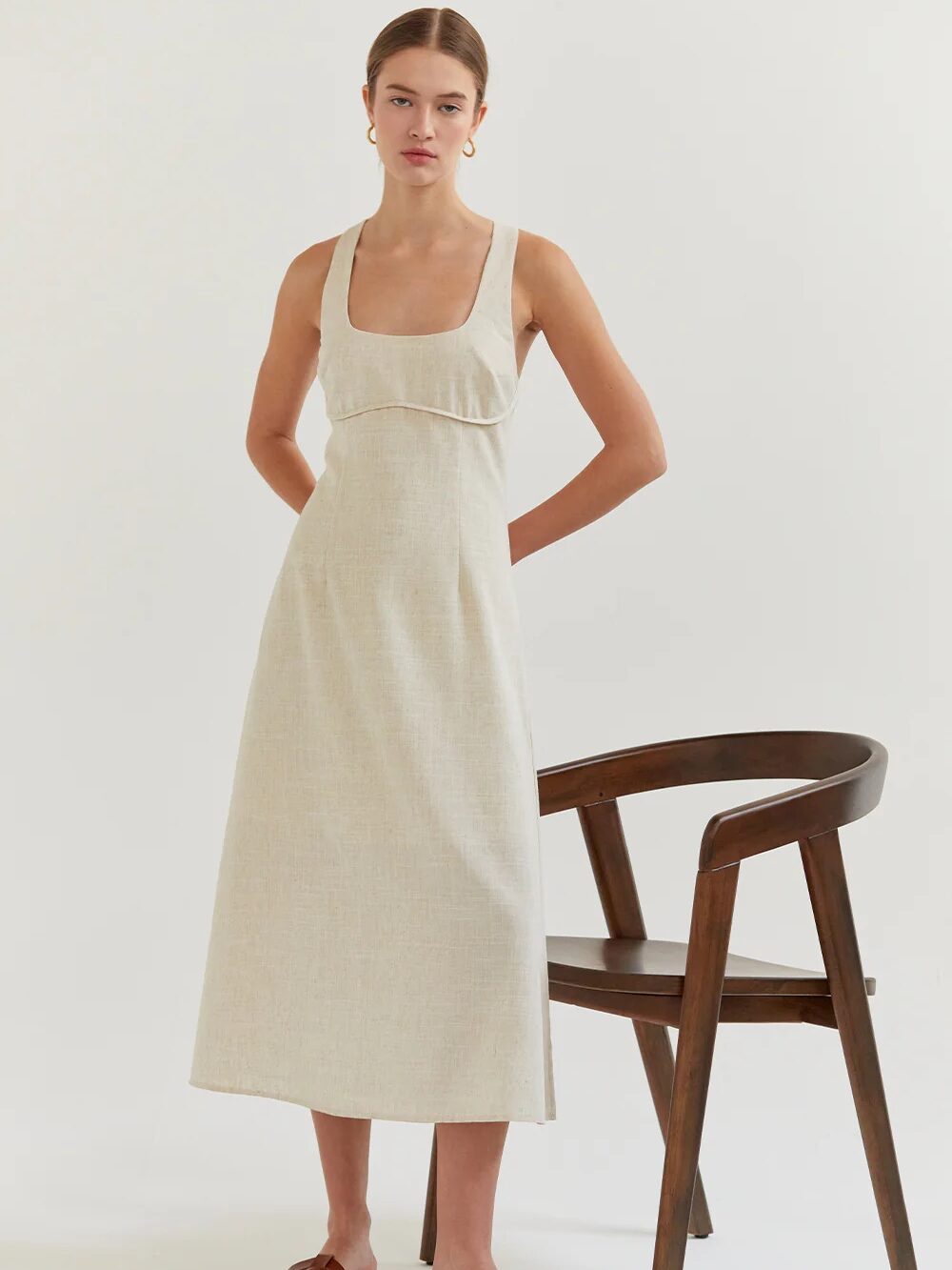 A young woman wearing a beige midi sundress stands next to a dark wooden chair on a plain background.