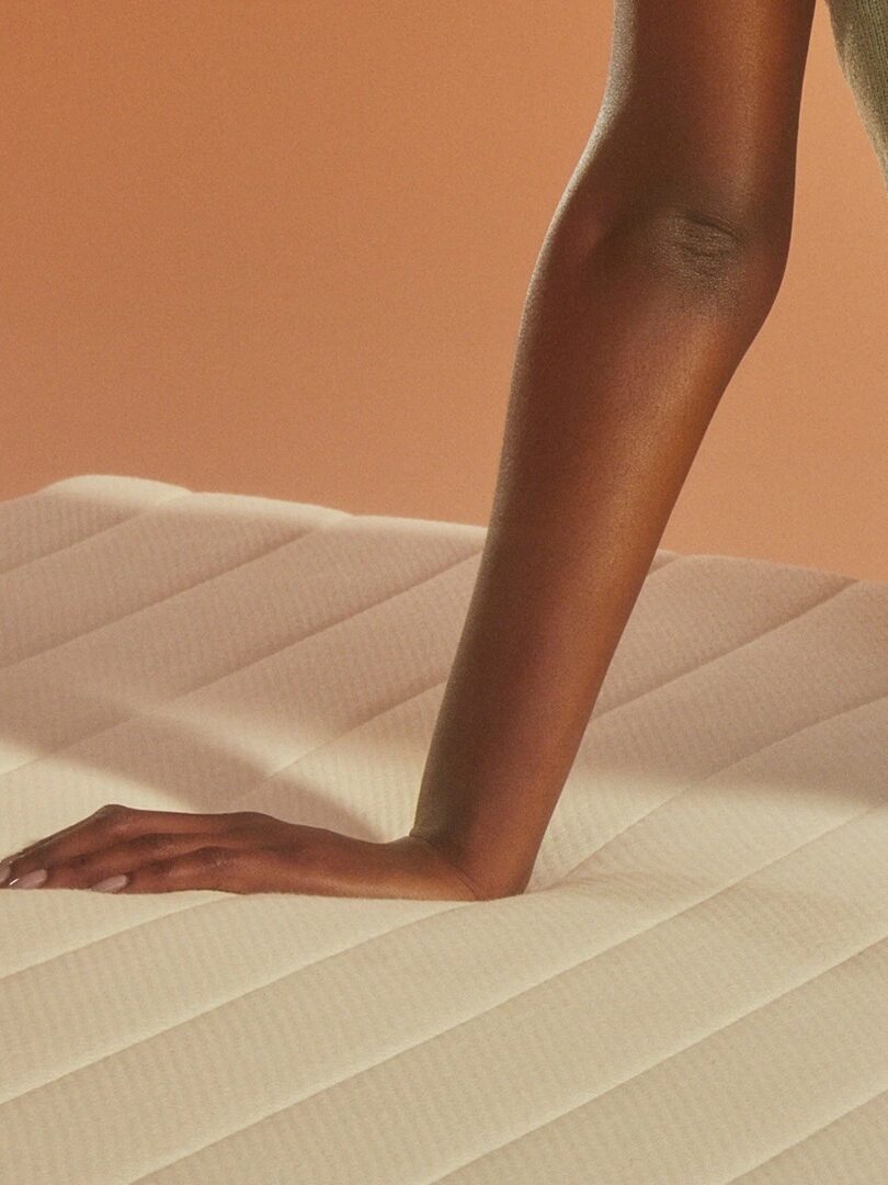 Person's hand and arm on a quilted mattress with a peach-colored background.